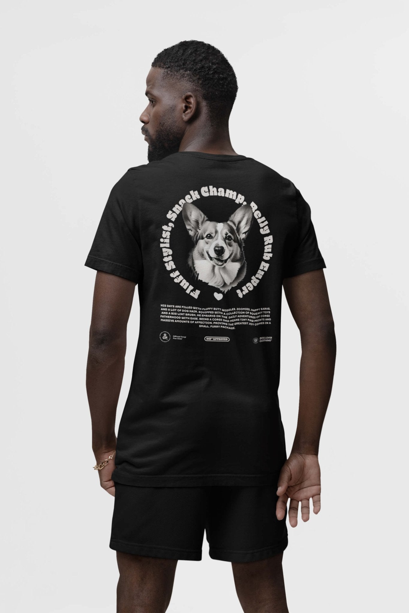 A man wearing black shorts and a Corgi Dad Ultimate Tribute T-Shirt from Dogloverclothing.com, featuring a dog in a circular design with text on the back, poses against a plain white background. Ideal for dog lovers or proud Corgi dads.