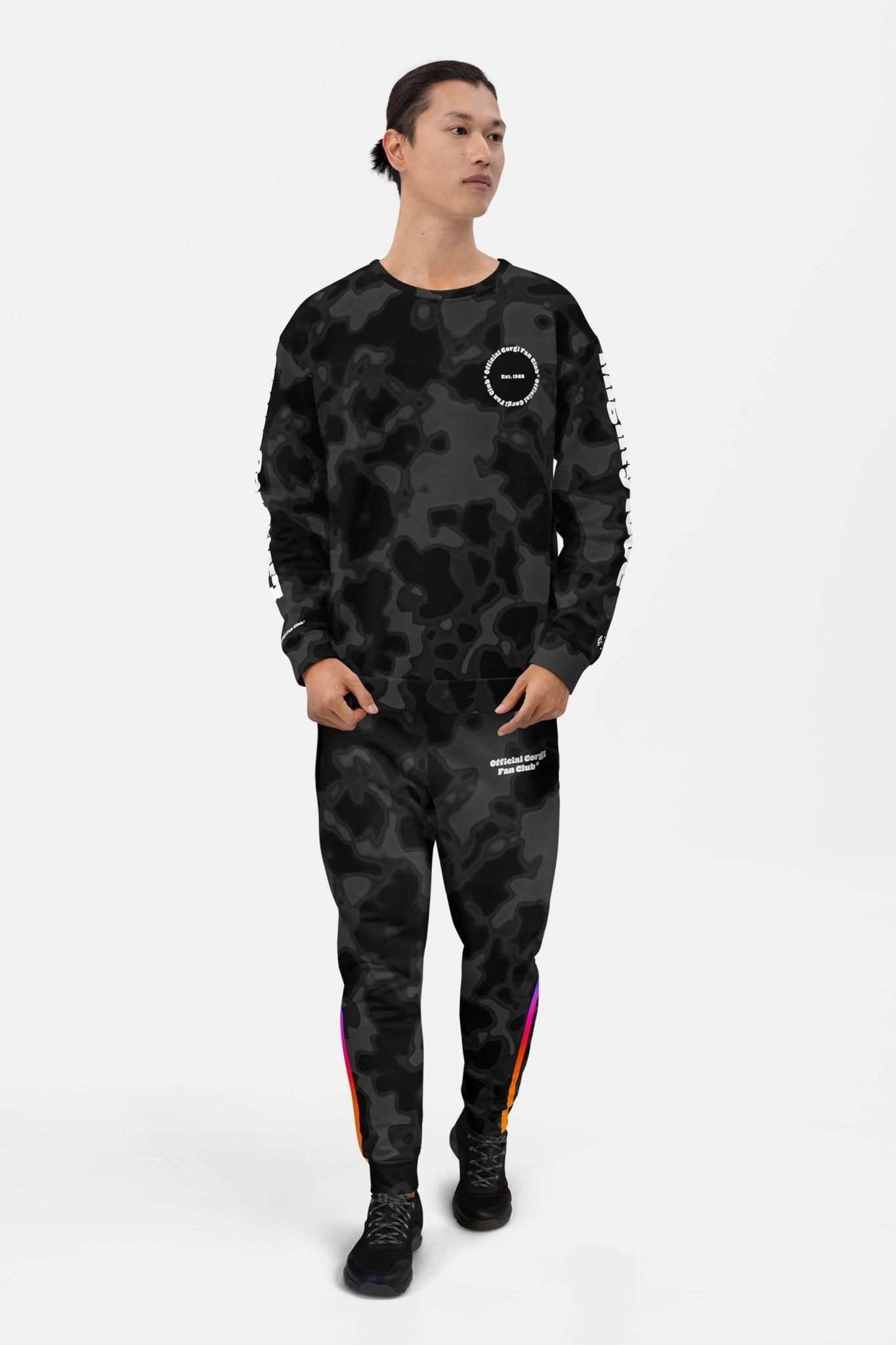 A person stands against a white background in the Official Corgi Fan Club© Elite Camo Sweatshirt and matching pants from Dogloverclothing.com, featuring a black and gray camo pattern, white text, colorful ankle stripe, and black sneakers.