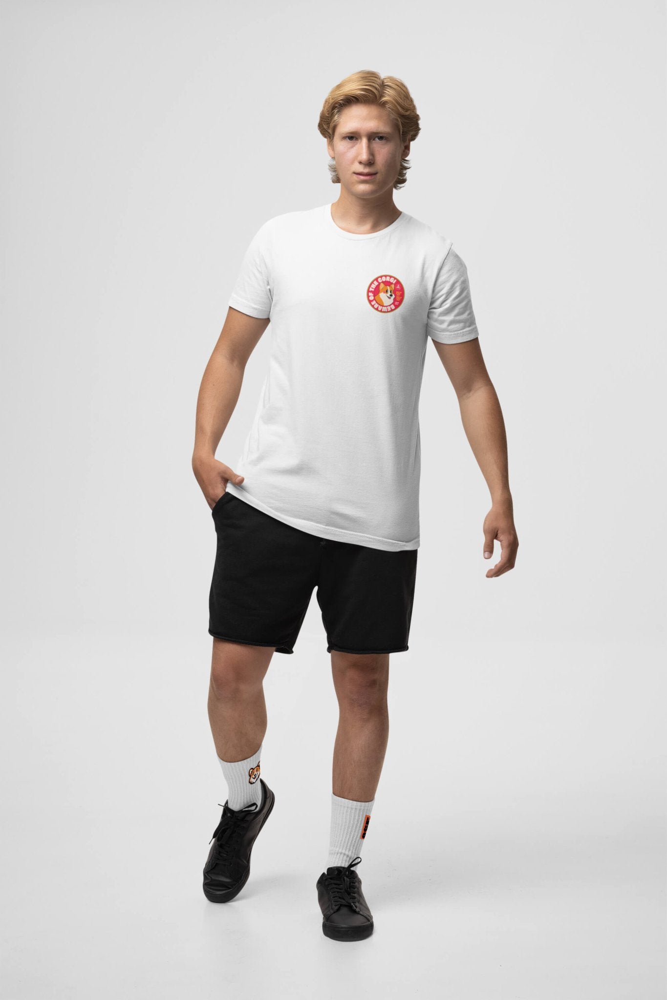 A person wearing a Beware of the Corgi Supreme unisex T-shirt from Dogloverclothing.com and black shorts is standing in a neutral pose against a plain white background. They have short blond hair, with super comfy white socks and black shoes completing the look.