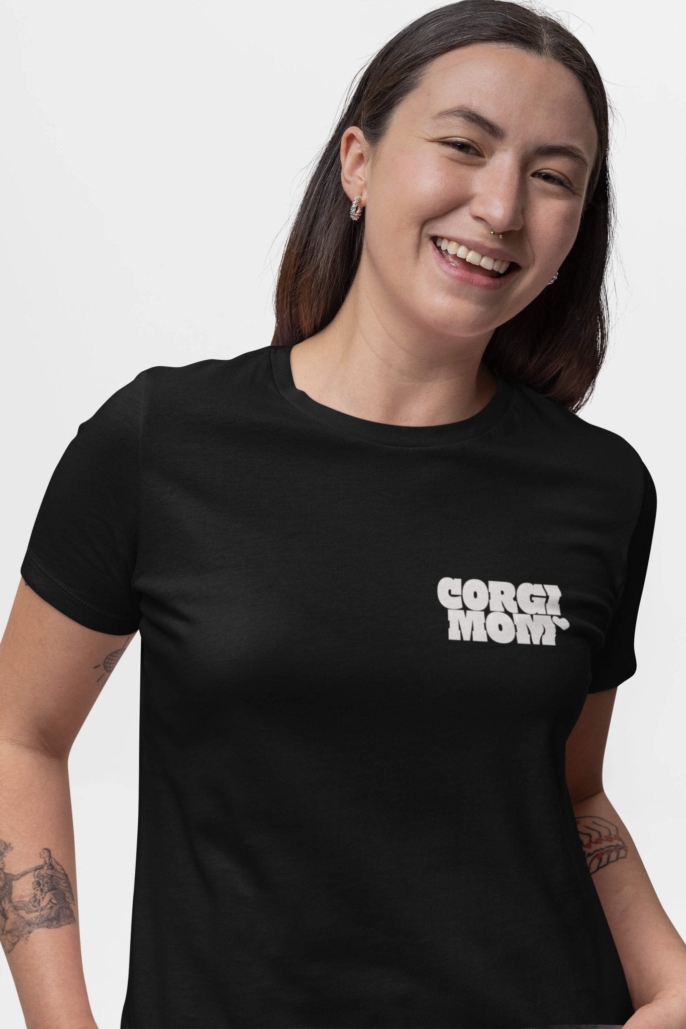 A person smiles in a Corgi Mom Ultimate Tribute T-Shirt from Dogloverclothing.com, crafted from black premium fabric featuring bold chest text. With long dark hair and arm tattoos against a plain white background, its the perfect blend of style for dog lovers.