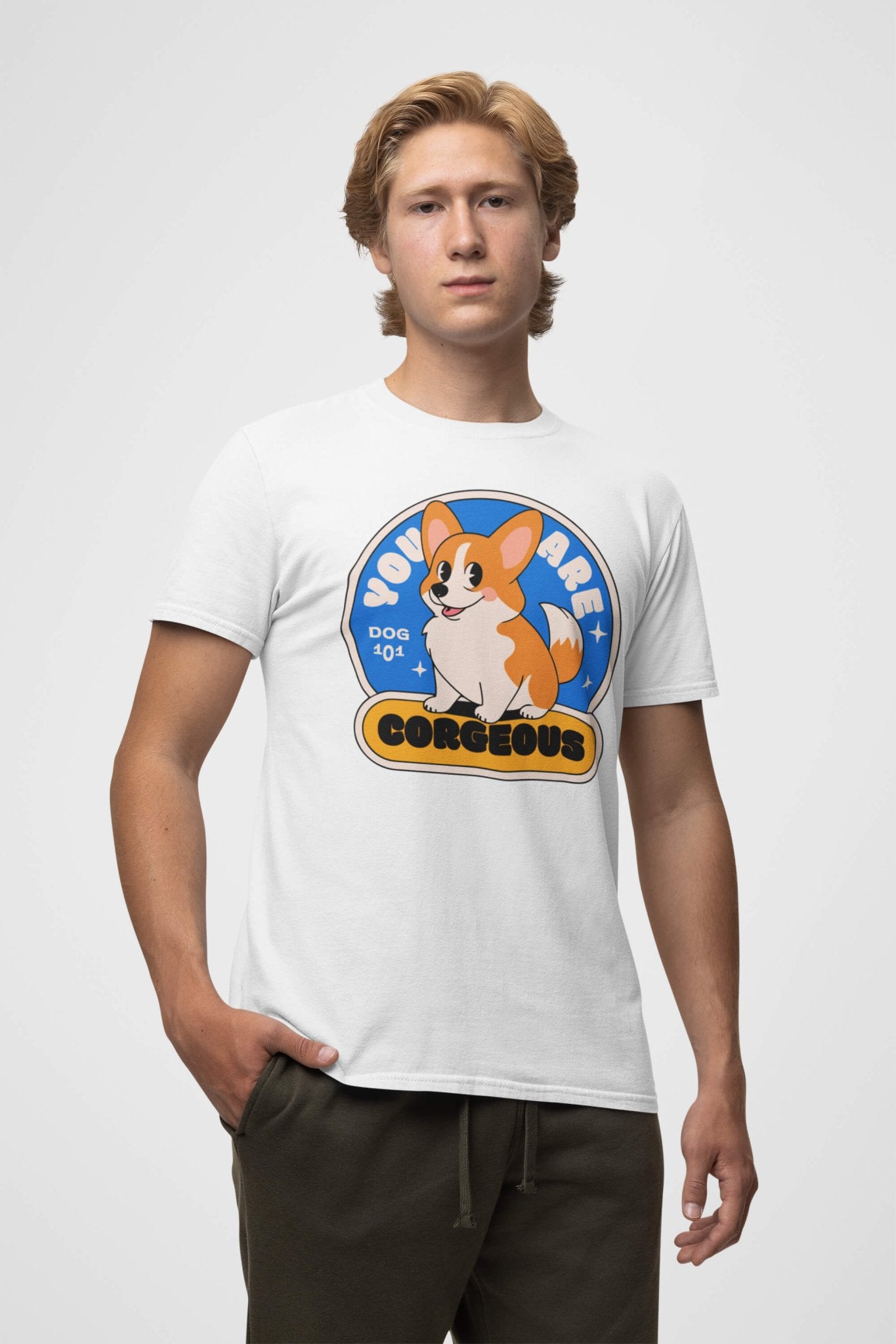 A person with light brown hair wears a Dogloverclothing.com You are Corgeous Supreme T-Shirt featuring a cartoon corgi and the text YOU ARE CORGEOUS and DOG 404. They stand with one hand in their pocket against a plain background—its perfect for dog lovers.