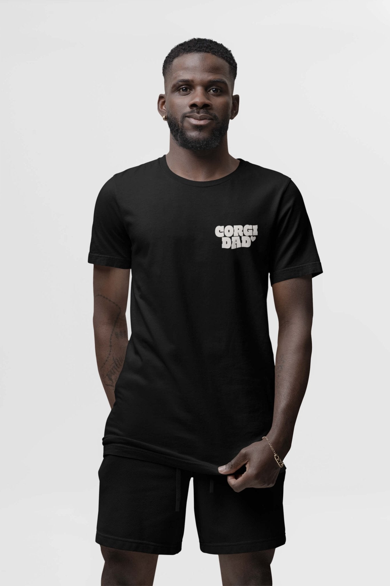 A man stands against a plain background wearing the black Corgi Dad Ultimate Tribute T-Shirt from Dogloverclothing.com, complemented by matching black shorts. Sporting a slight smile and short beard, he exudes a casual vibe with one hand resting on his shorts. Perfect for dog lovers!.