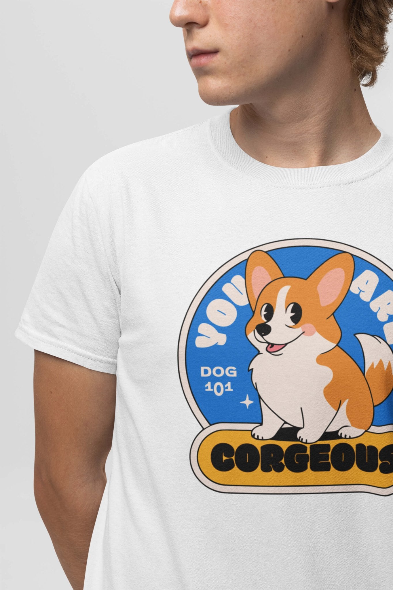 A person in a You are Corgeous Supreme T-Shirt by Dogloverclothing.com, featuring a cheerful cartoon Corgi with stars and the text You Are Corgeous and Dog 101, looks to the side against a gray background. Perfect for dog lovers!.