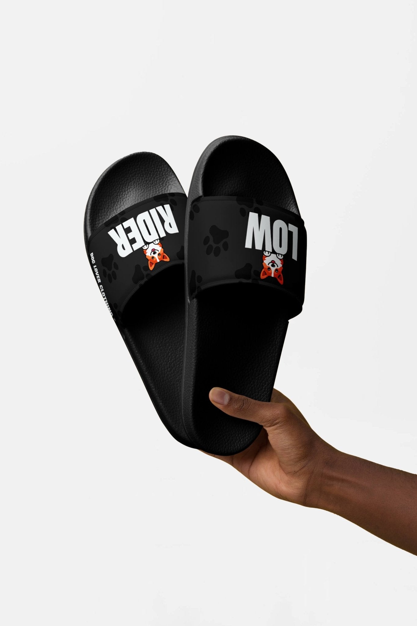 A hand displays Mighty Corgi Rider Slides from Dogloverclothing.com. The black slides feature paw prints, “LOW” and “RIDER” straps with a cute corgi, crafted from durable materials and standing out against a white background.