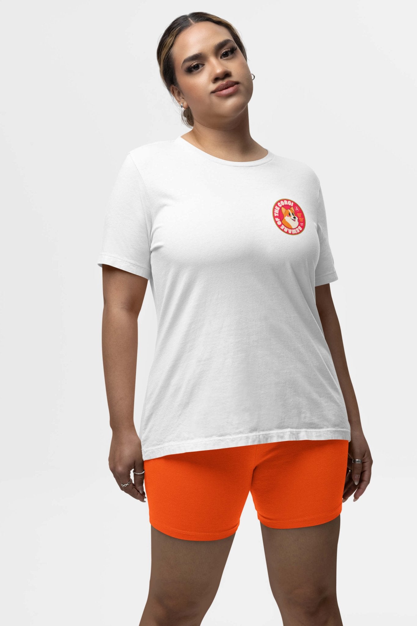 A person in a Beware of the Corgi Supreme Unisex T-Shirt by Dogloverclothing.com, featuring a circular emblem for Corgi enthusiasts, paired with bright orange shorts stands against a plain backdrop. The comfy fabric complements their relaxed posture and neutral expression perfectly.