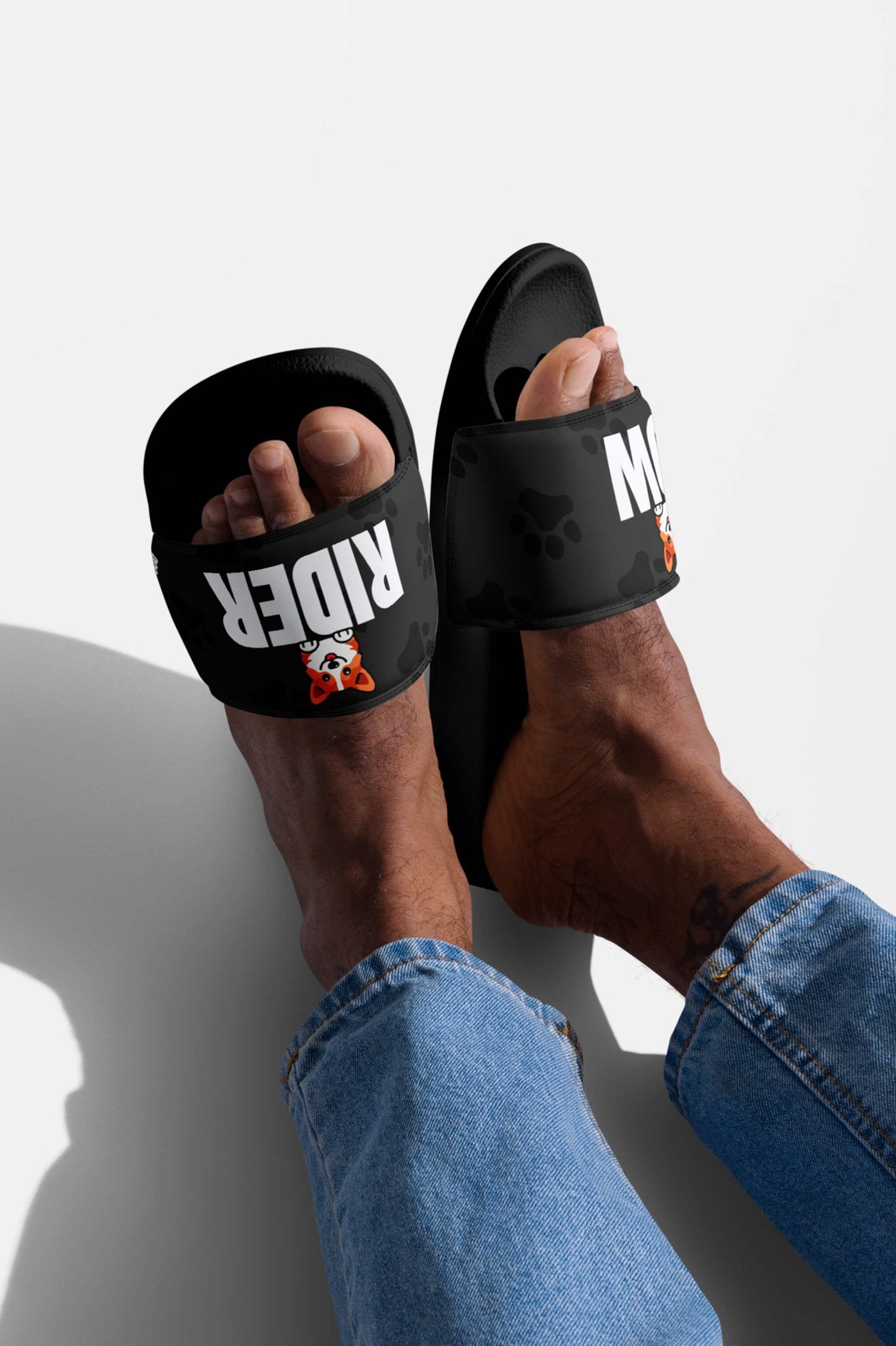 A person in blue jeans stands on a white surface, wearing Dogloverclothing.coms Mighty Corgi Rider Slides—black sandals with RIDER in bold white letters and a small orange cartoon—showcasing their stylish, comfy footwear.
