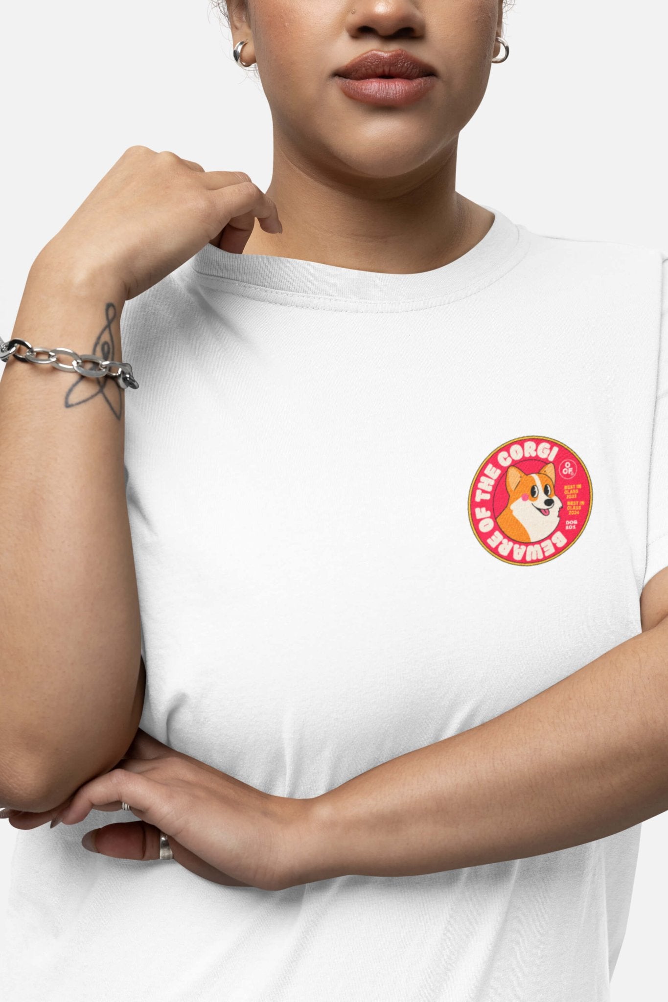A person sports the Beware of the Corgi Supreme Unisex T-Shirt from Dogloverclothing.com, featuring a round corgi patch and Corgi Life. Ideal for corgi enthusiasts, it offers comfy fabric. With short hair, light makeup, and silver jewelry, their hand rests gently near the shoulder.