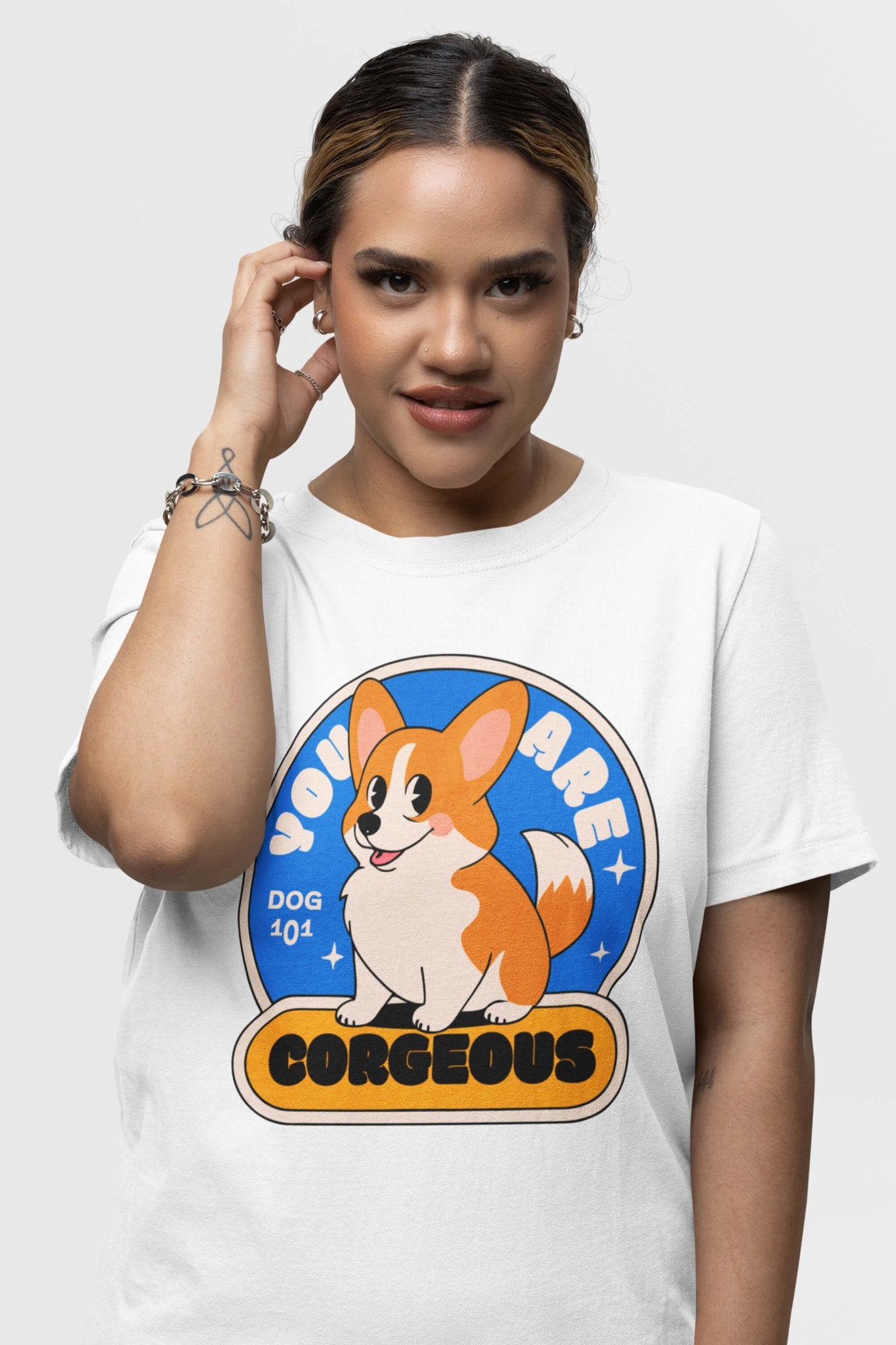A person with a casual hairstyle wears Dogloverclothing.coms You Are Corgeous Supreme T-Shirt, featuring a cartoon corgi and text. Complemented by a silver bracelet, they pose against a plain background, making it ideal for dog enthusiasts.