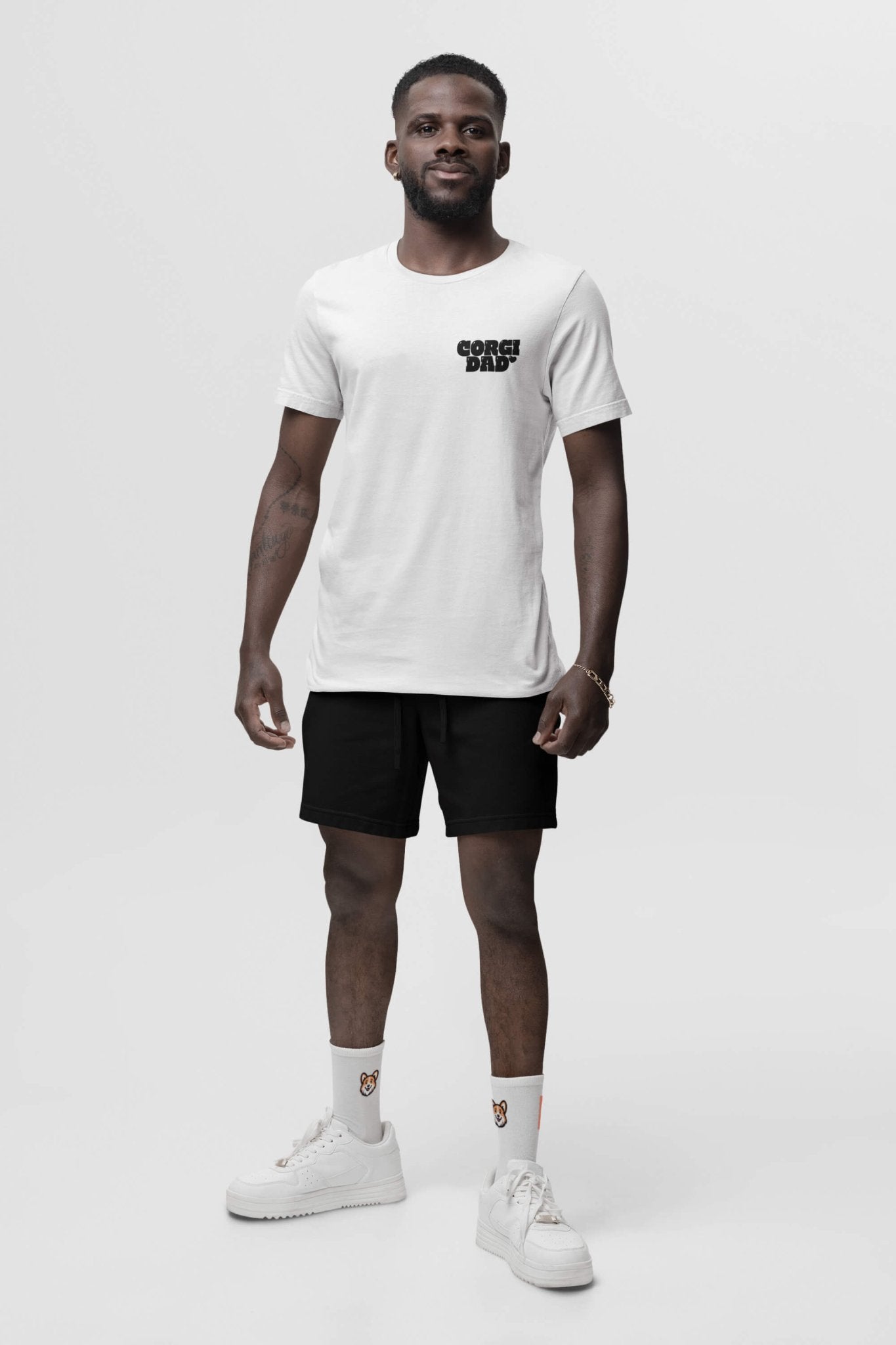 Against a plain white background, a smiling person proudly wears the Corgi Dad Ultimate Tribute T-Shirt from Dogloverclothing.com, crafted from premium cotton. They pair it with black shorts, patterned white socks, and white sneakers. Their short hair and beard contribute to their relaxed style.