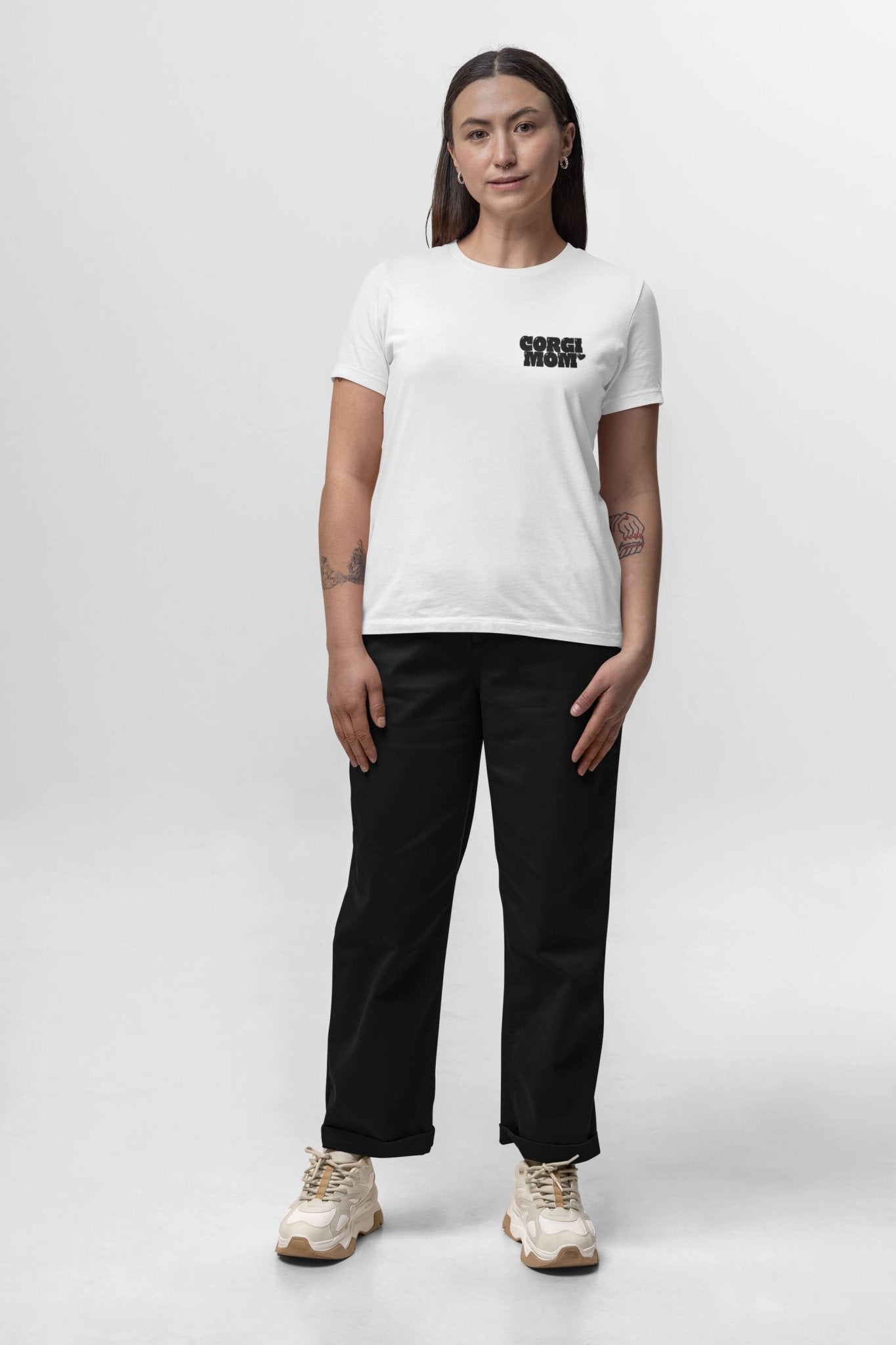 Facing forward against a plain background, the person wears a white Corgi Mom Ultimate Tribute T-Shirt from Dogloverclothing.com featuring black text on the chest and made of premium fabric. They pair it with black pants and white sneakers. Their long hair cascades past tattooed forearms, perfect for dog lovers.