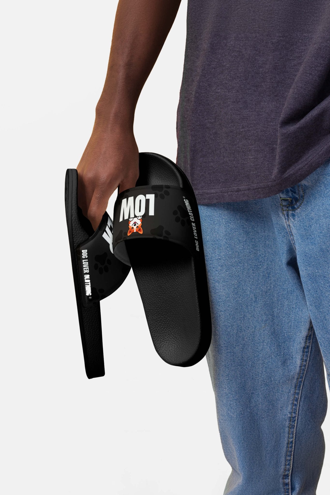 A person in blue jeans and a dark shirt holds the Mighty Corgi Rider Slides from Dogloverclothing.com, black slides with LOW and a cartoon dog on the straps—ideal for fans of comfortable footwear.