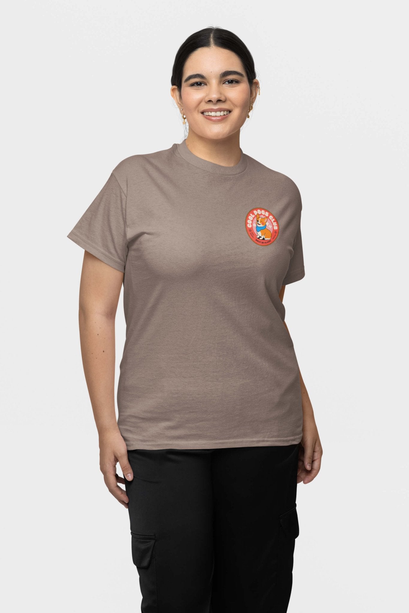 A person with dark hair is smiling, wearing a beige Cool Dogs Club unisex hype t-shirt from Dogloverclothing.com with a red circular logo, paired with black pants against a plain white background, perfect for dog lovers.