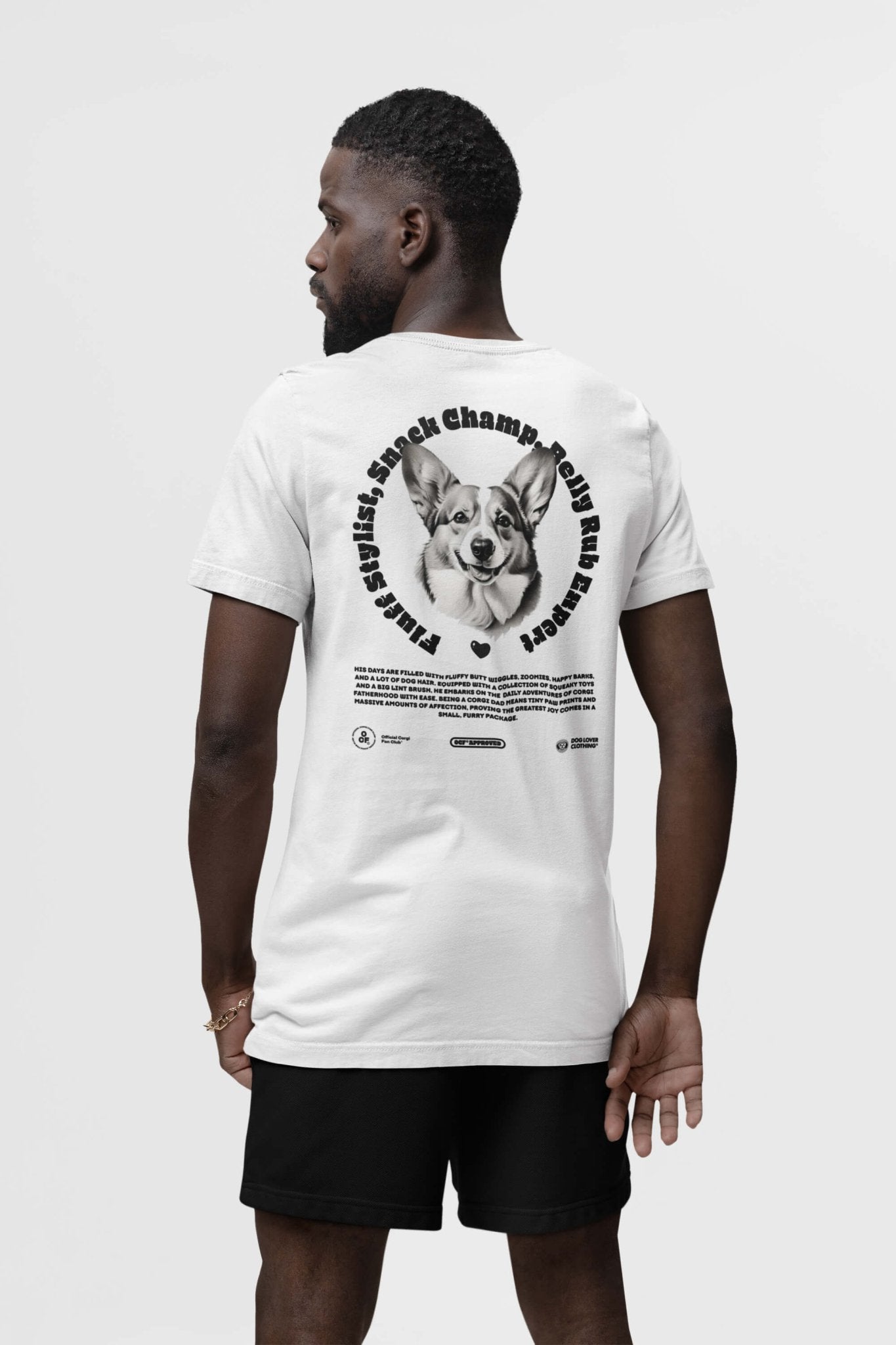 A man in a white Corgi Dad Ultimate Tribute T-Shirt from Dogloverclothing.com flaunts a smiling corgi graphic encircled by Snack Champion Club and Paws & Jaws, making it perfect for dog lovers. The unisex shirt pairs stylishly with black shorts, ideal for any canine enthusiast.