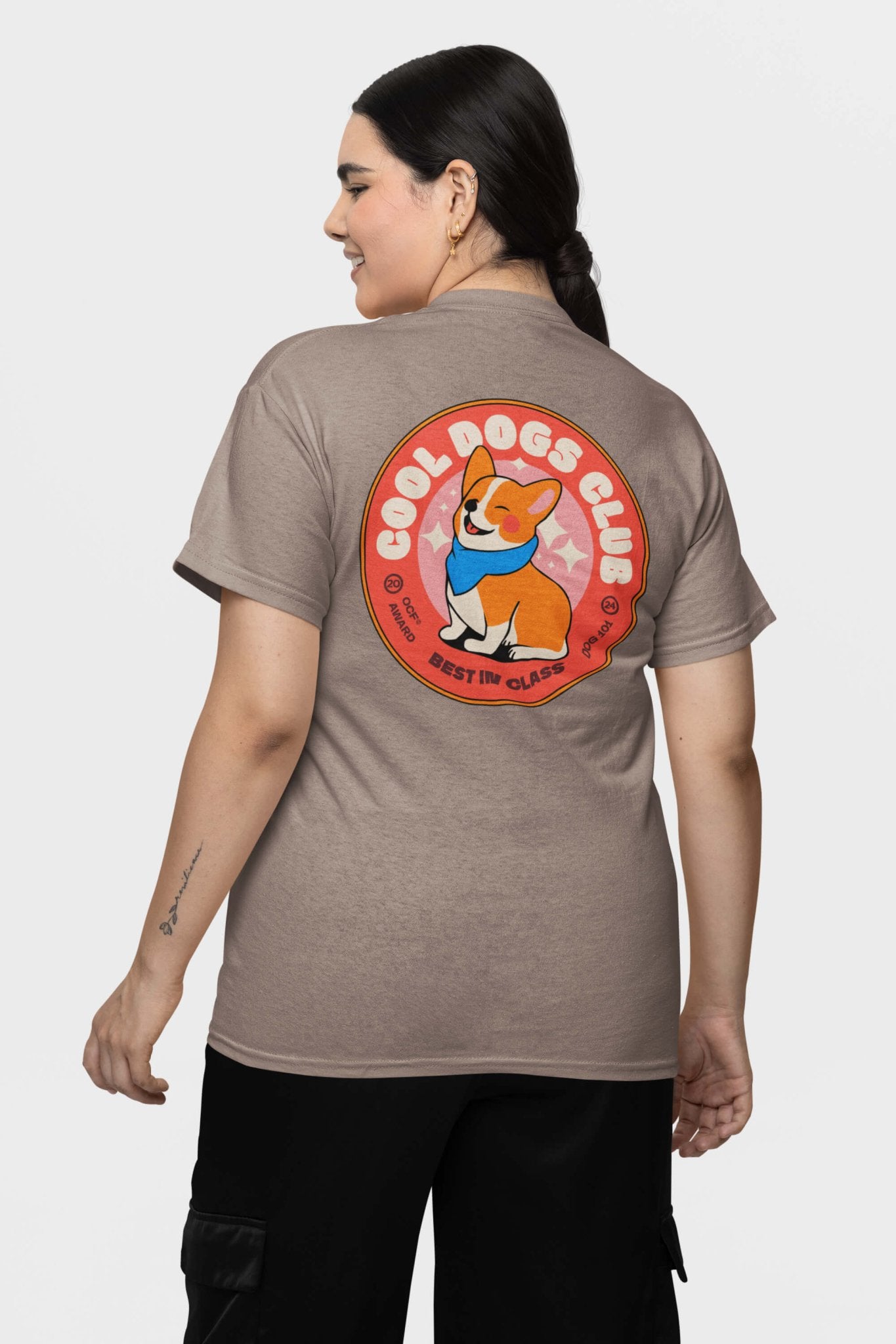 A woman models a Cool Dogs Club Unisex Hype T-Shirt from Dogloverclothing.com, featuring a vibrant cartoon dog graphic and Best in Class text on the back. Perfect for dog lovers and Corgi t-shirt fans, she smiles with her dark hair tied back as she faces away from the camera.