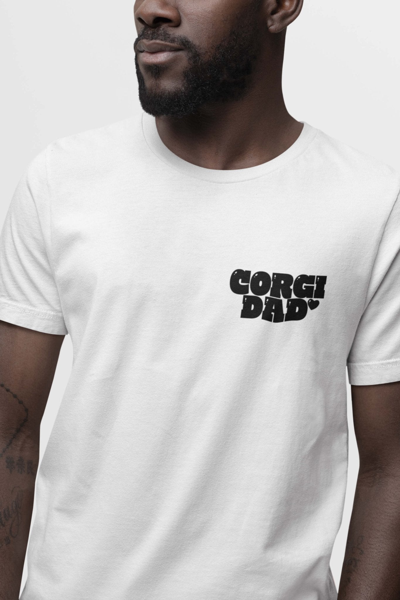 A tattooed man confidently wears the Corgi Dad Ultimate Tribute T-Shirt from Dogloverclothing.com, with bold black text on his chest. Perfect for dog lovers, this look stands out against a neutral backdrop.