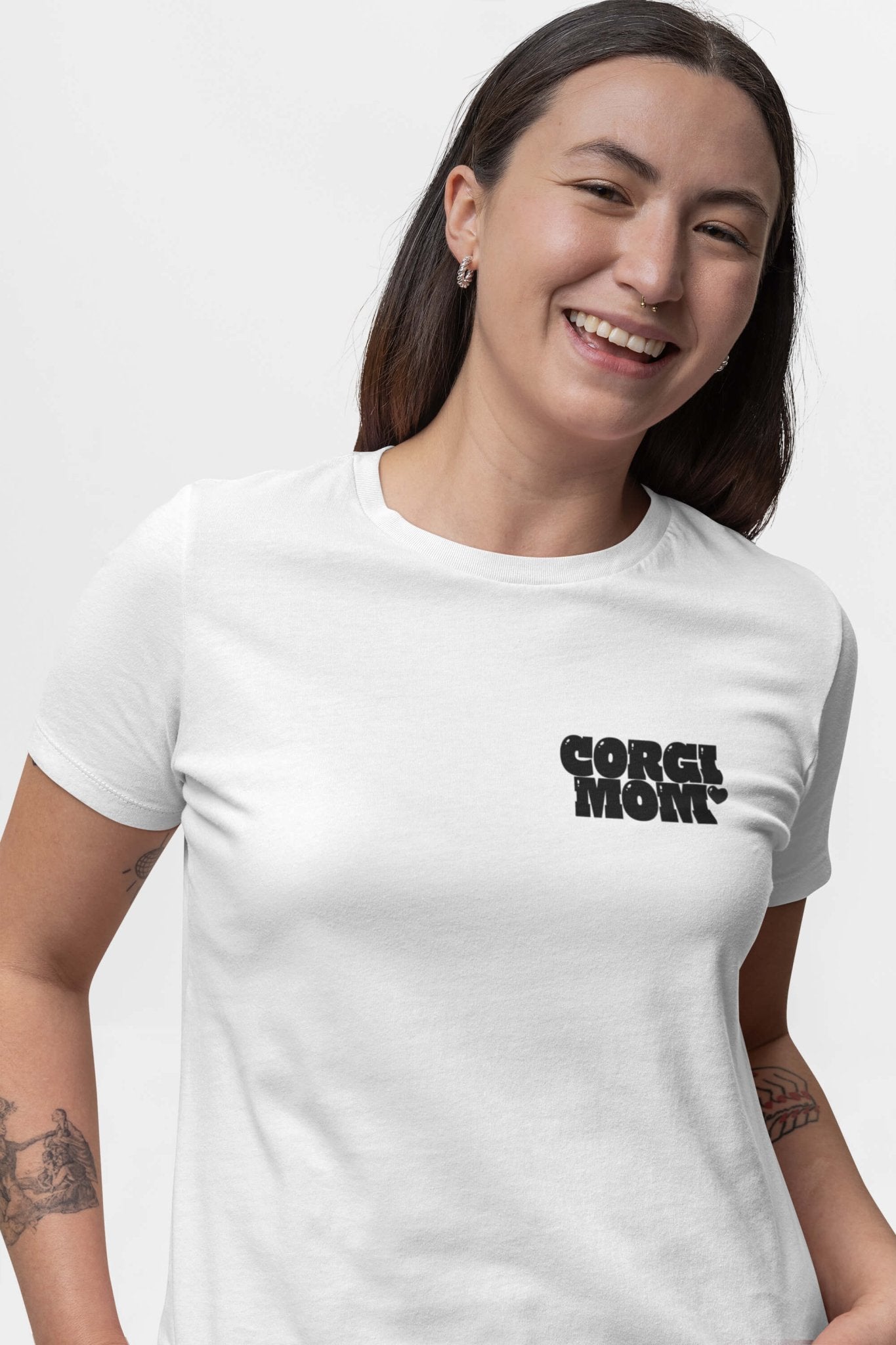 A person smiles, wearing a Corgi Mom Ultimate Tribute T-Shirt from Dogloverclothing.com. They have long hair, small earrings, and tattoos on their arms, standing against a plain white background—a great pick for dog lovers.