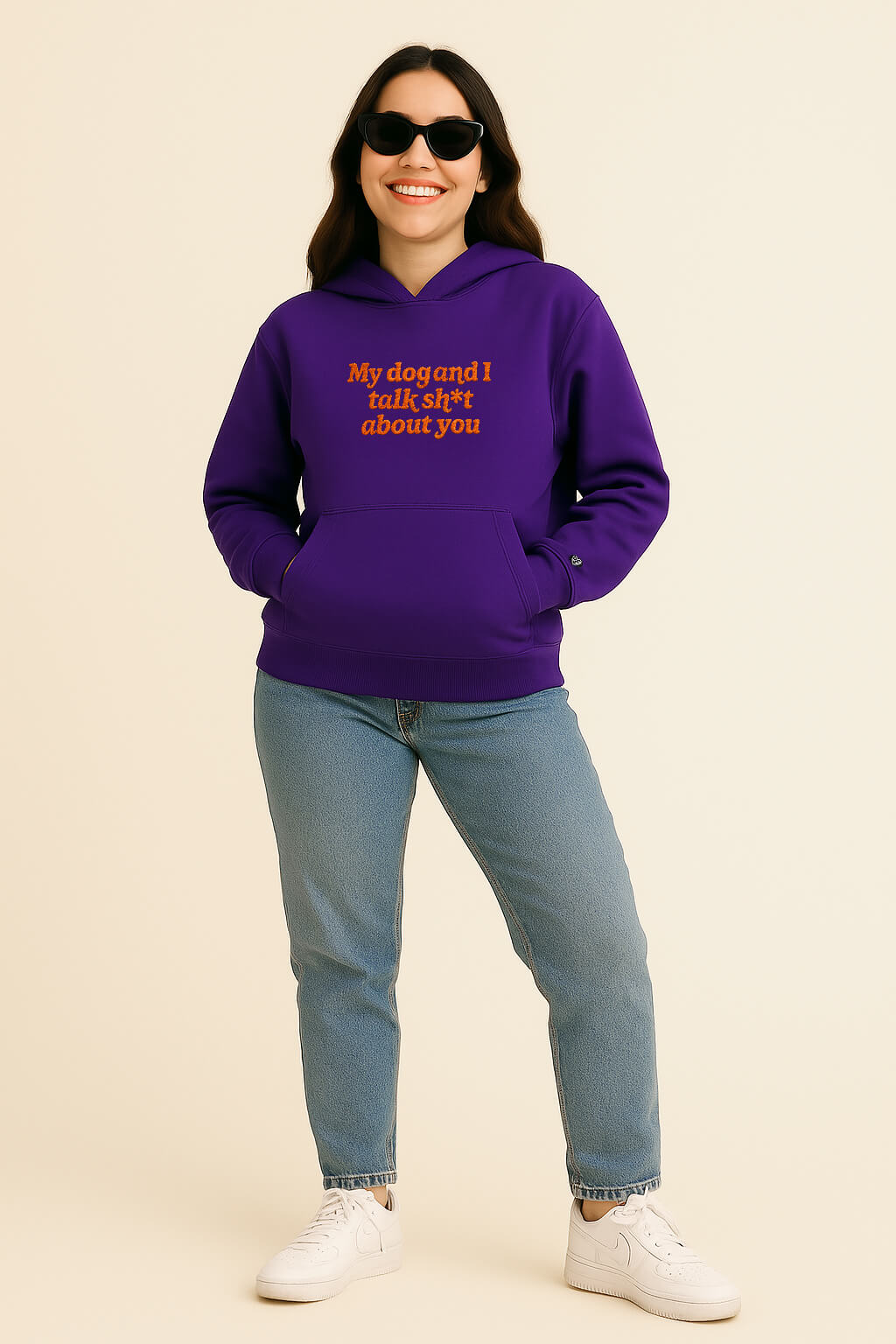 A smiling woman in sunglasses wears the Bold My Dog And I Talk Embroidered Hoodie by Dogloverclothing.com, blue jeans, and white sneakers, standing against a plain cream background with her hands in her hoodie pockets.