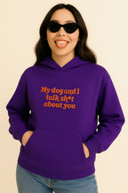 A woman in black cat-eye sunglasses smiles, tongue out and hands in pockets, wearing the Bold My Dog And I Talk Embroidered Hoodie by Dogloverclothing.com—a purple premium hoodie with “My dog and I talk sh*t about you” embroidered in orange.