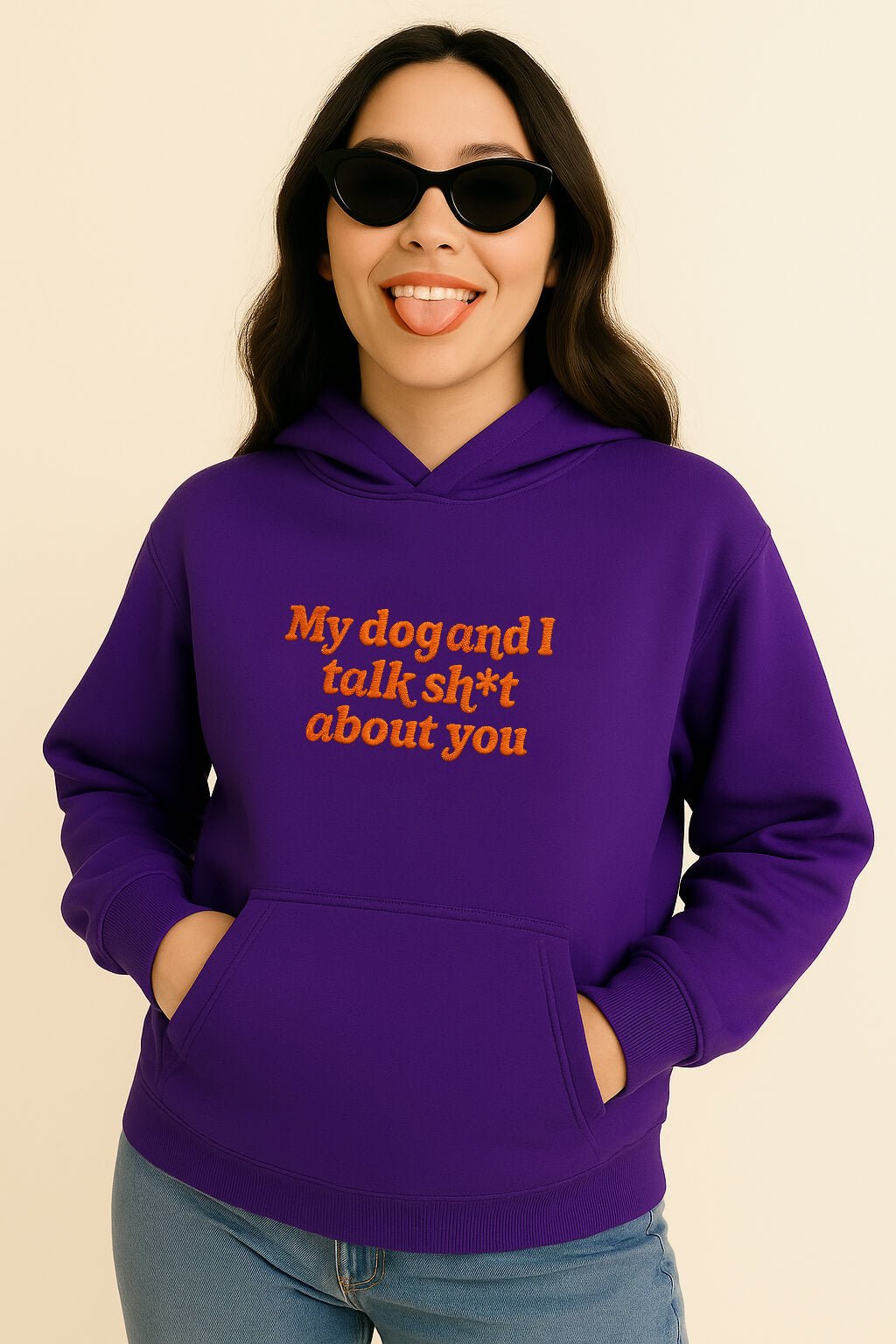 A woman in black cat-eye sunglasses smiles, tongue out and hands in pockets, wearing the Bold My Dog And I Talk Embroidered Hoodie by Dogloverclothing.com—a purple premium hoodie with “My dog and I talk sh*t about you” embroidered in orange.