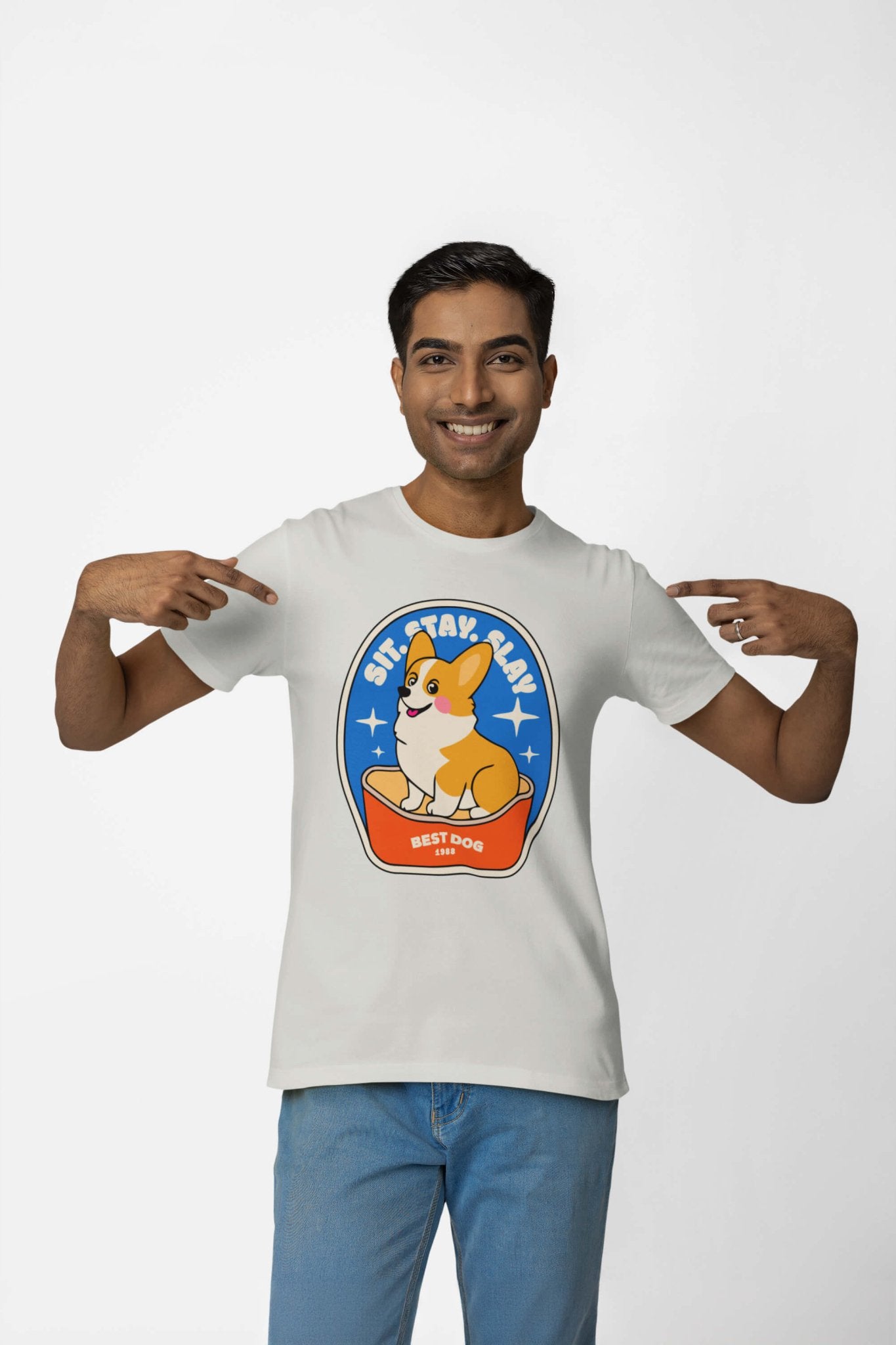 A person wears a Best Dog Supreme Tribute Unisex T-Shirt from Dogloverclothing.com. It features a premium cotton light-colored tee with an oval corgi design and Sit, Stay, Slay and Best Dog text. Smiling, they point at this must-have for dog lovers.