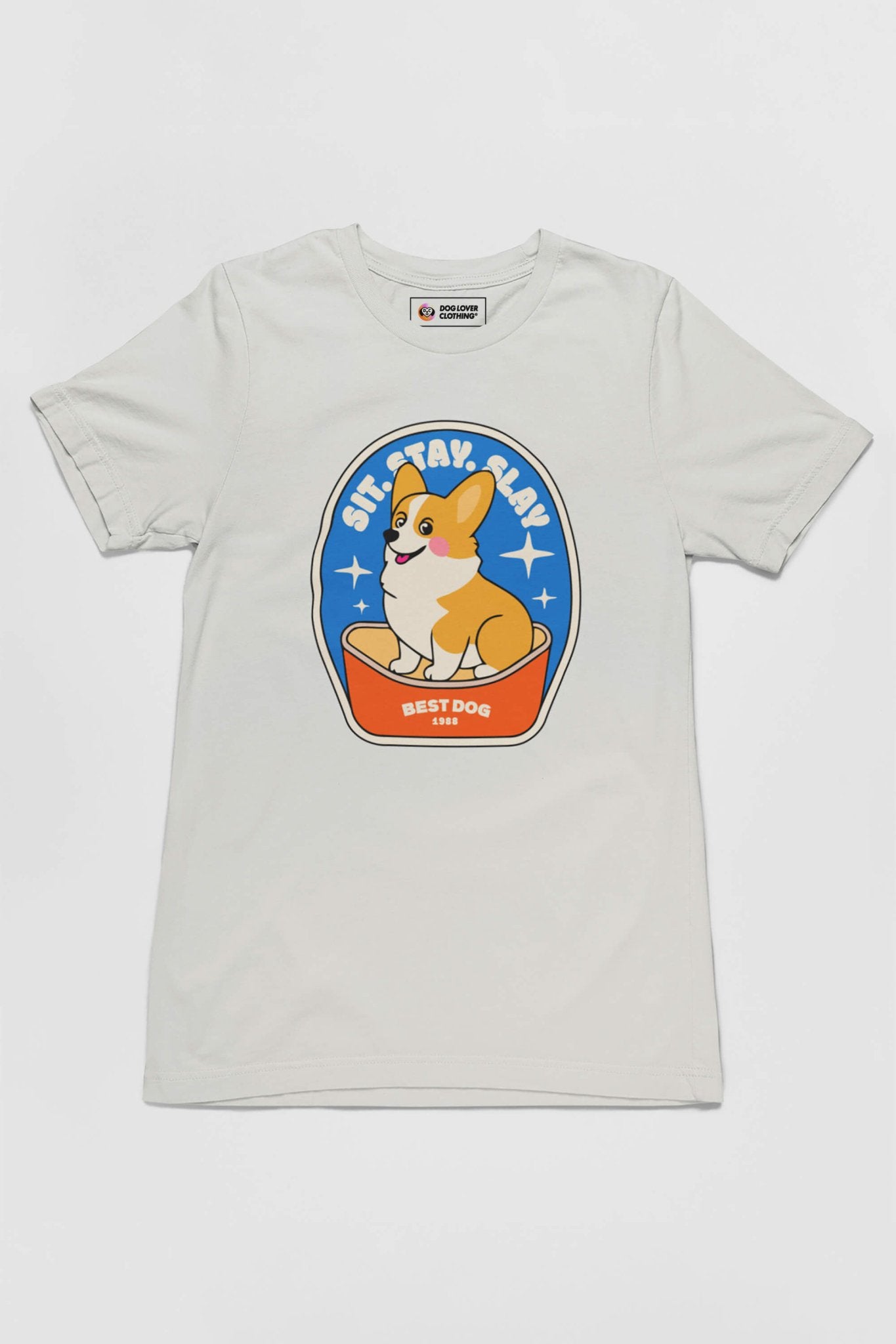 The Best Dog Supreme Tribute Unisex T-Shirt from Dogloverclothing.com features a vivid cartoon Corgi perched in a bowl with Sit, Stay, Lay above and Best Dog below, on premium cotton with a bright blue background—perfect for dog lovers.