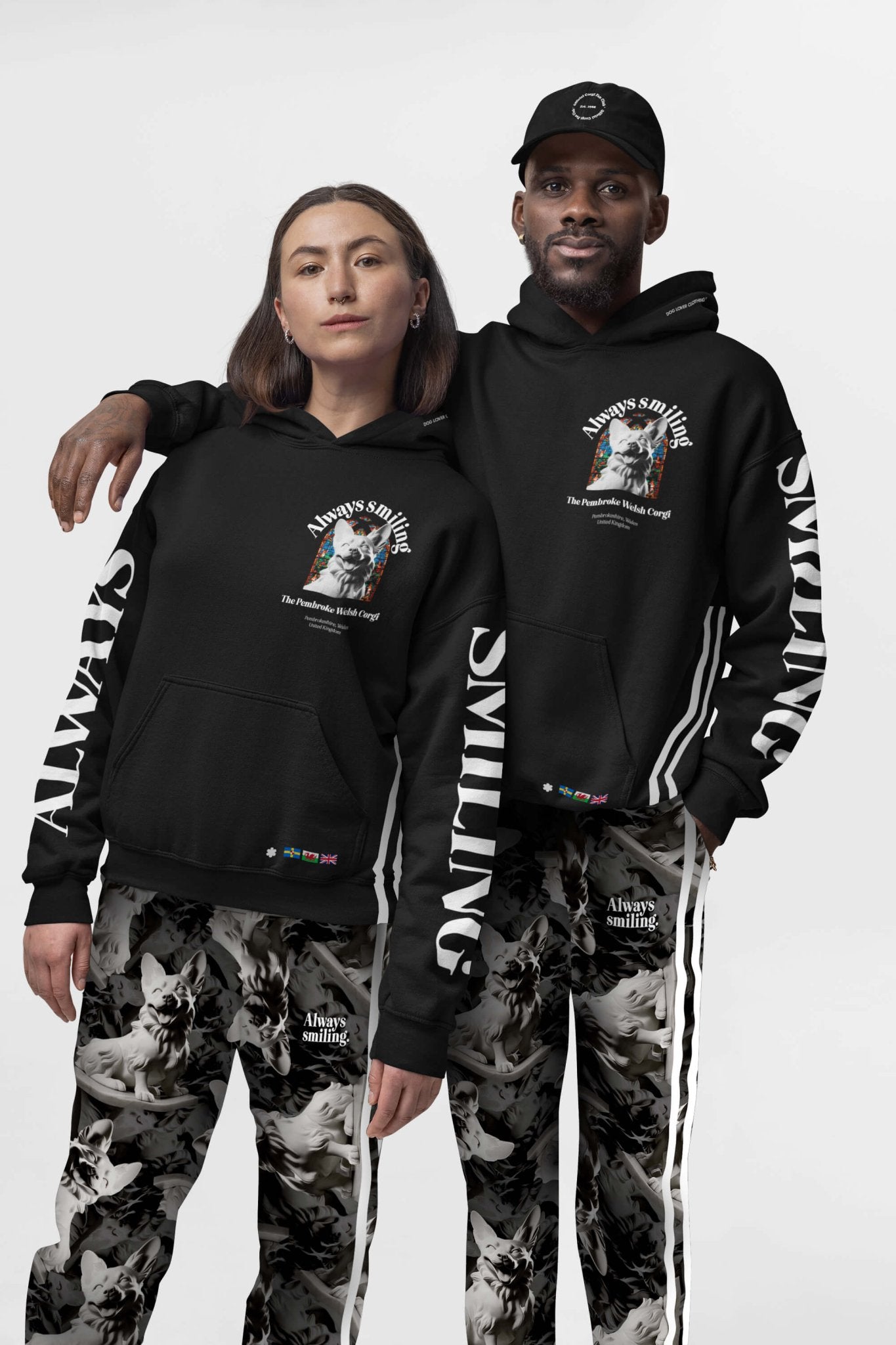In matching Corgi Supreme Tribute Hoodies by Dogloverclothing.com, two people pose with Always Smiling graphics. One wears a black cap, both sport abstract patterned pants. The sleeves read ALWAYS and SMILING, set against a neutral backdrop—ideal apparel for dog lovers.