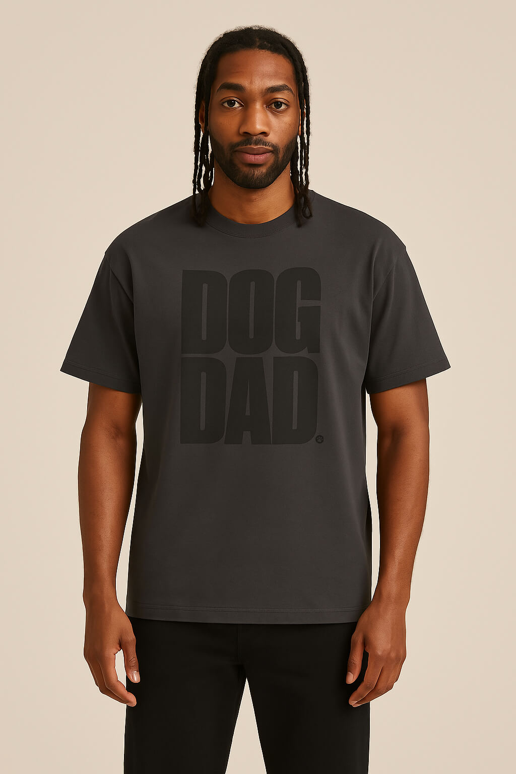 A man with long braids wears the Oversized Dog Dad T-Shirt by Dogloverclothing.com, featuring DOG DAD. in bold on a black, durable cotton tee. He stands against a plain beige background facing forward with a neutral expression.