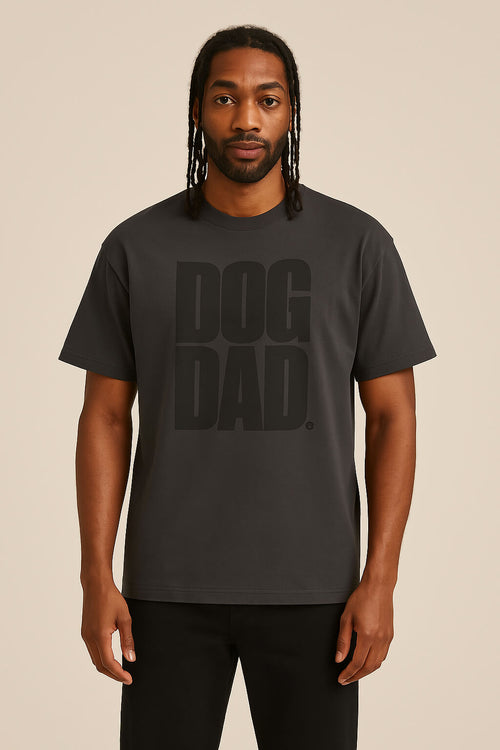 A man with long braids wears the Oversized Dog Dad T-Shirt by Dogloverclothing.com, featuring DOG DAD. in bold on a black, durable cotton tee. He stands against a plain beige background facing forward with a neutral expression.