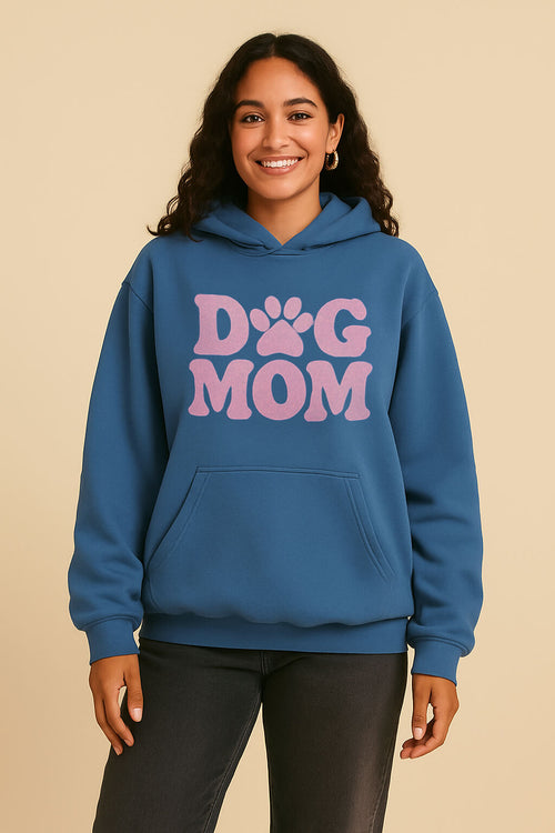 A smiling person wears the Dogloverclothing.com Dog Mom Hoodie, an ultra-soft blue hoodie with large pink “Dog Mom” text and a playful paw-print, standing against a plain beige background.