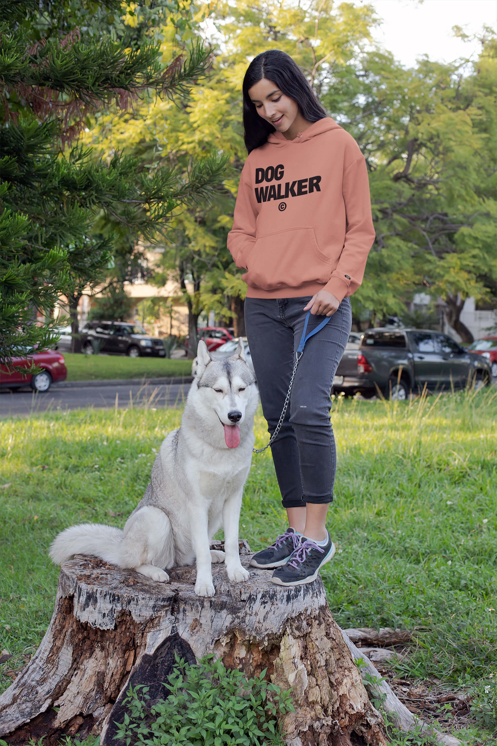 A woman wearing a Unisex Dog Walker© Premium Hoodie from Dogloverclothing.com stands on a tree stump with a gray and white husky. The leashed dog sits calmly, tongue out, surrounded by lush greenery and parked cars, creating an ideal setting for everyday adventures.