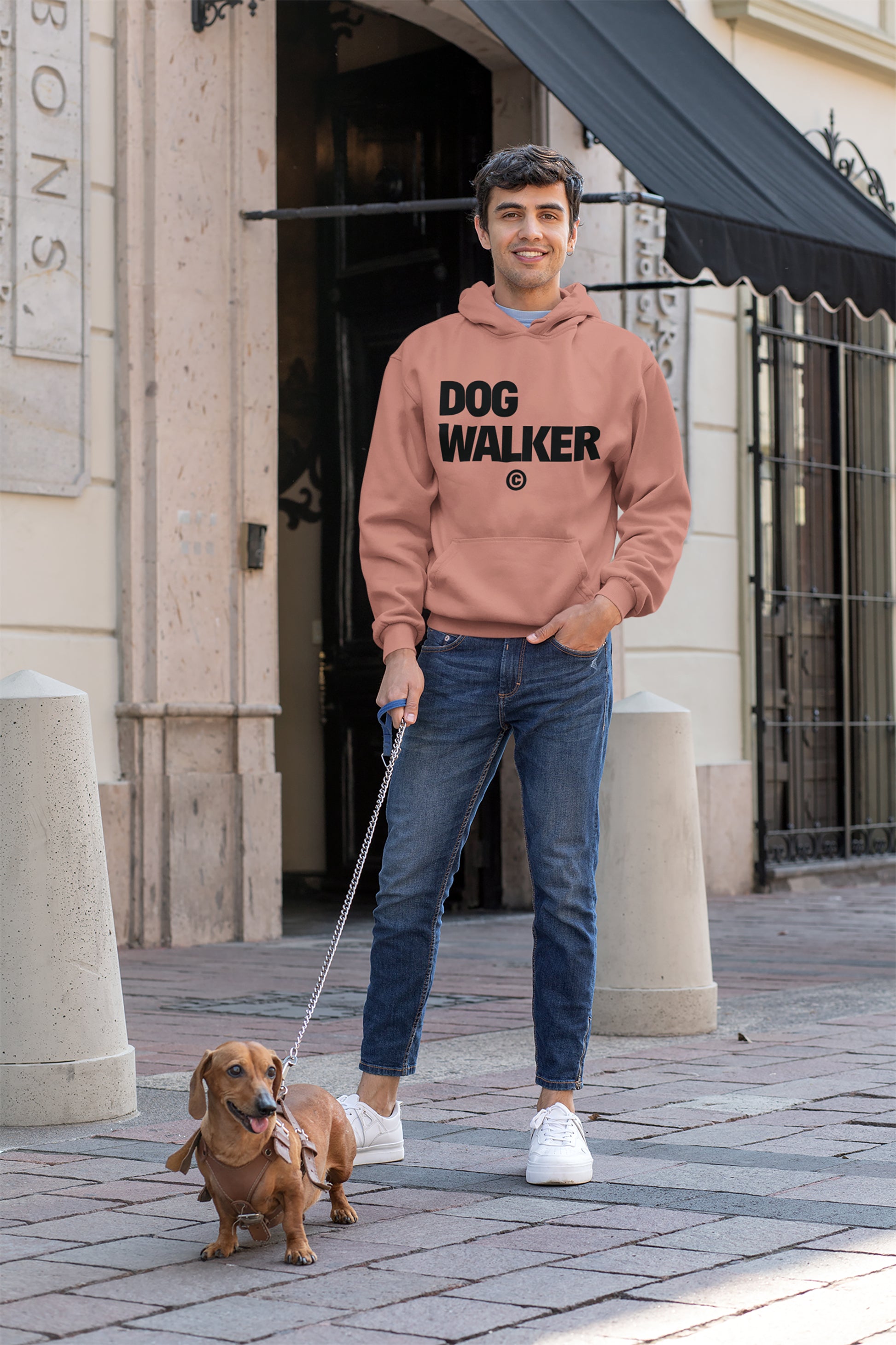 A person in a Dogloverclothing.com Unisex Dog Walker© Premium Hoodie and jeans smiles, holding a leash attached to a small brown dog, ready for everyday adventures on a sidewalk before large building windows.