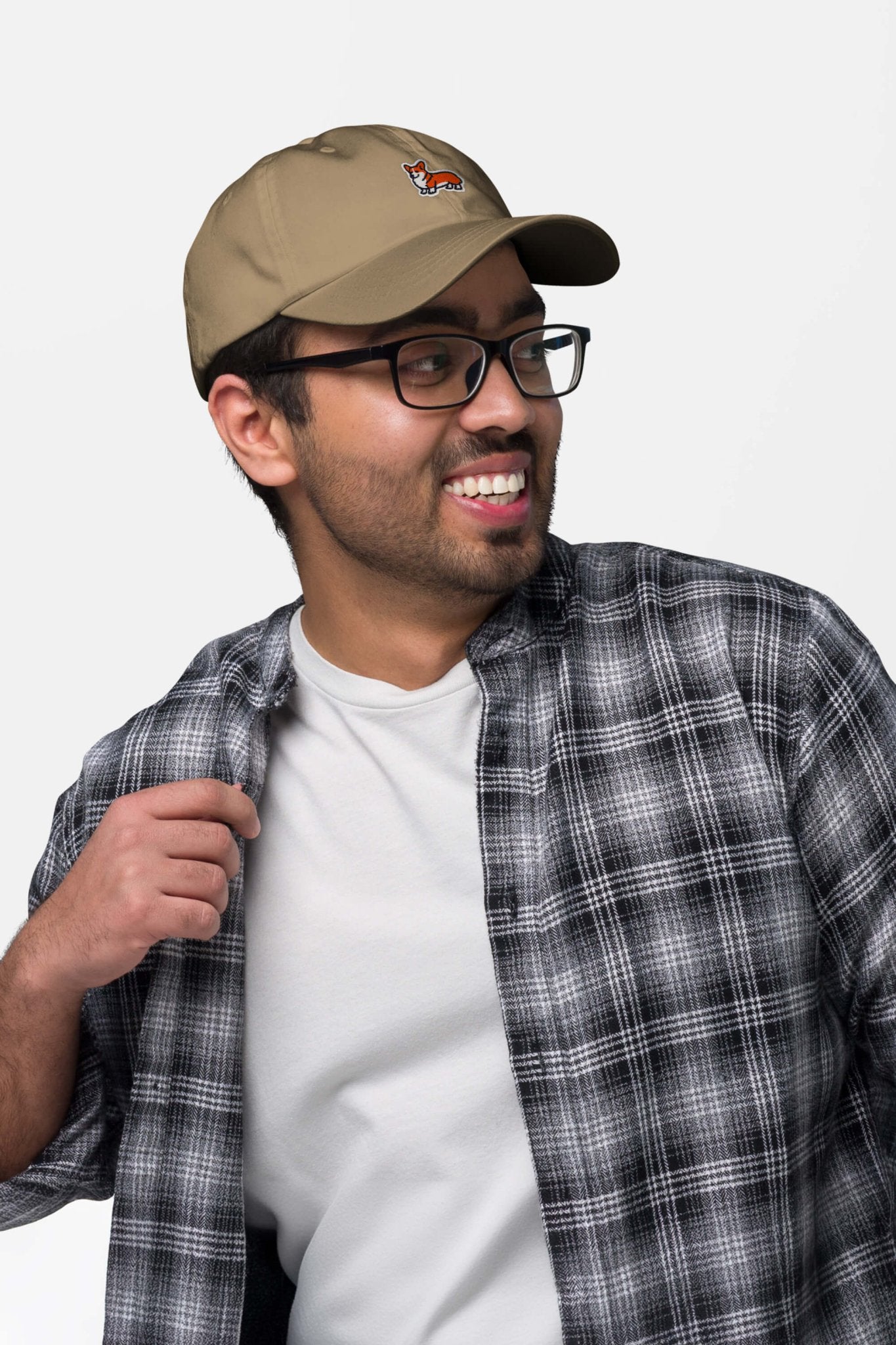 A man in glasses, a white tee, and a black-and-white plaid shirt smiles to the side while wearing the Iconic Corgi Cap from Dogloverclothing.com, showing off its small embroidered fox against a plain light background.