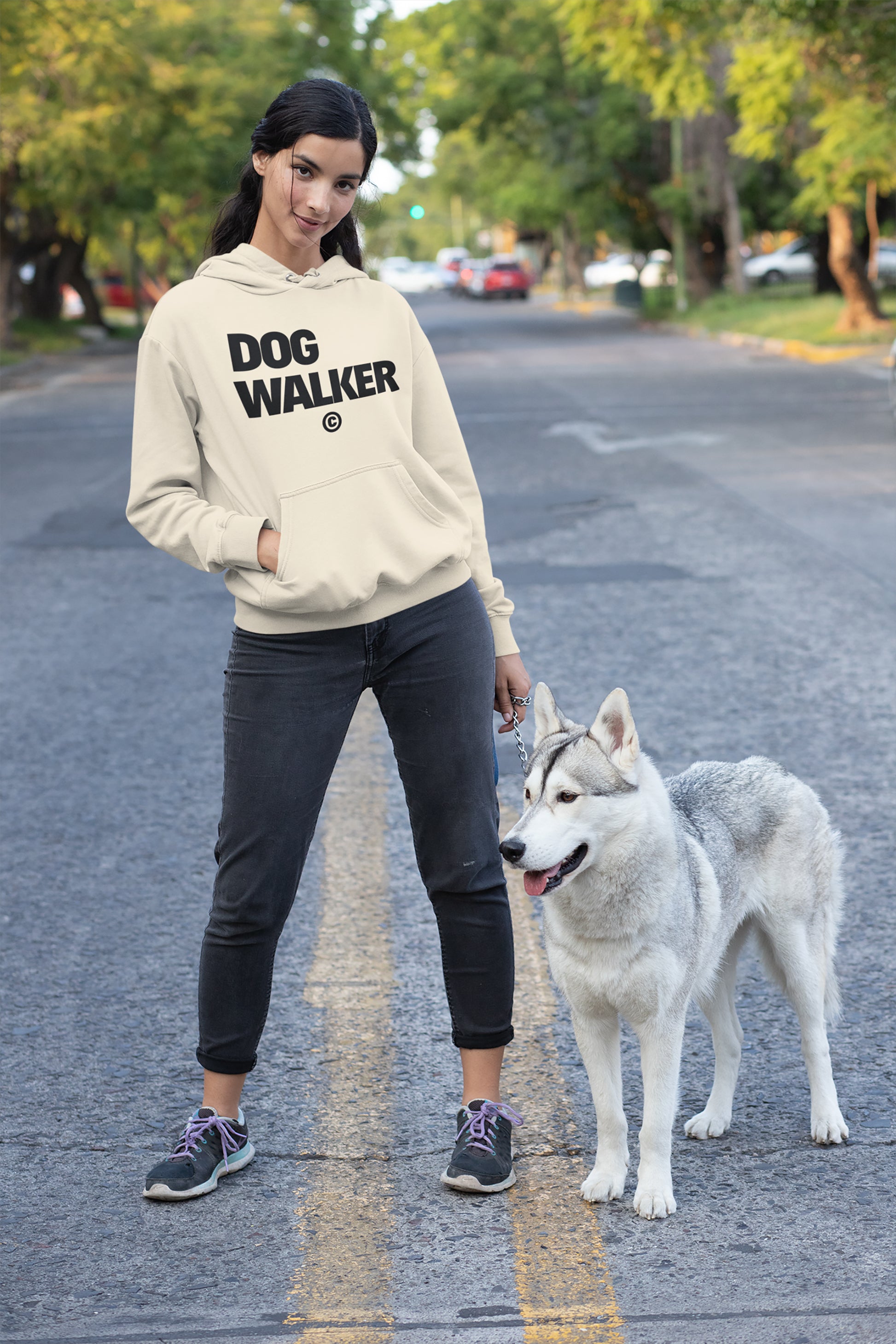 Confidently standing on a suburban street with their large white and gray dog on a leash, the person wears black pants and an Unisex Dog Walker© Premium Hoodie from Dogloverclothing.com. Trees and parked cars line the background under the sunny sky.