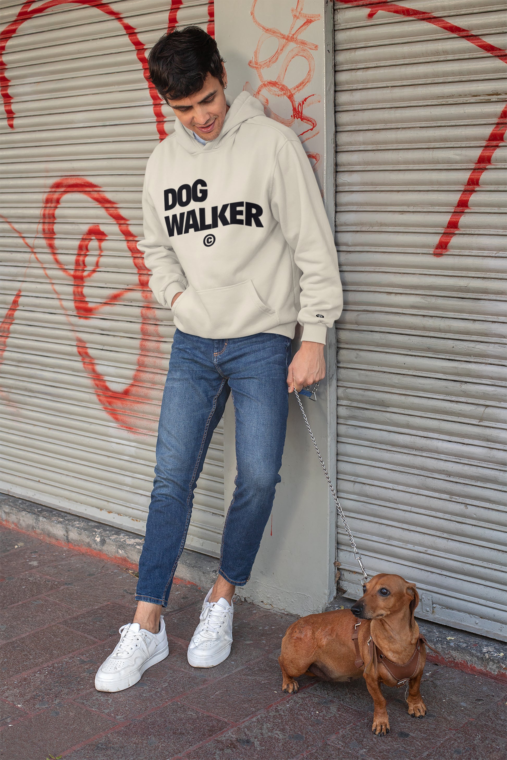 In front of a graffiti-covered shutter, someone wears a Unisex Dog Walker© Premium Hoodie in beige from Dogloverclothing.com, paired with blue jeans and white sneakers. They hold a leash attached to a small brown dog, set for everyday adventures.