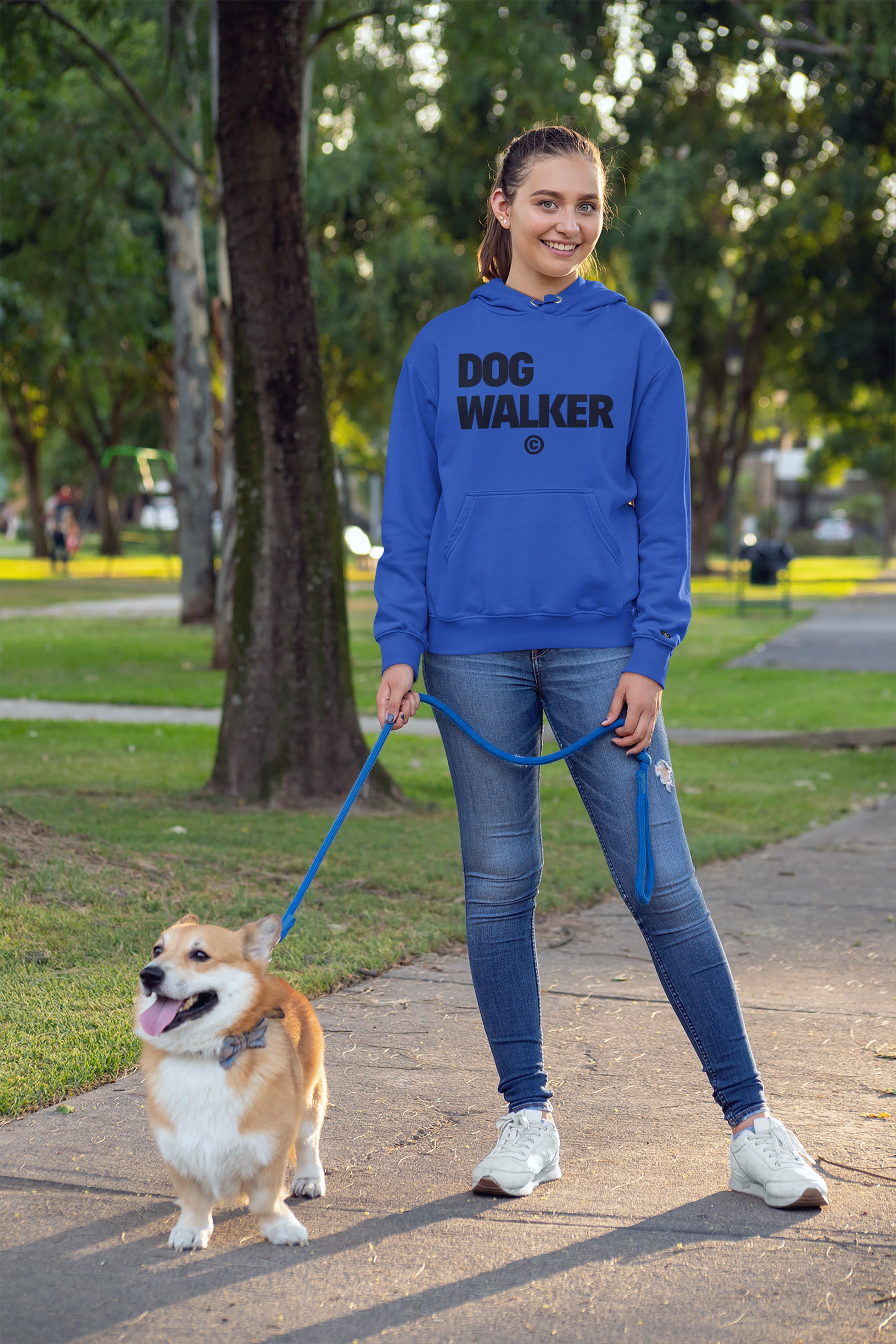 Wearing a Unisex Dog Walker© Premium Hoodie from Dogloverclothing.com, a person leads a corgi on an adventure through the park. The hoodie labeled DOG WALKER stands out as both smile amidst trees and winding paths.