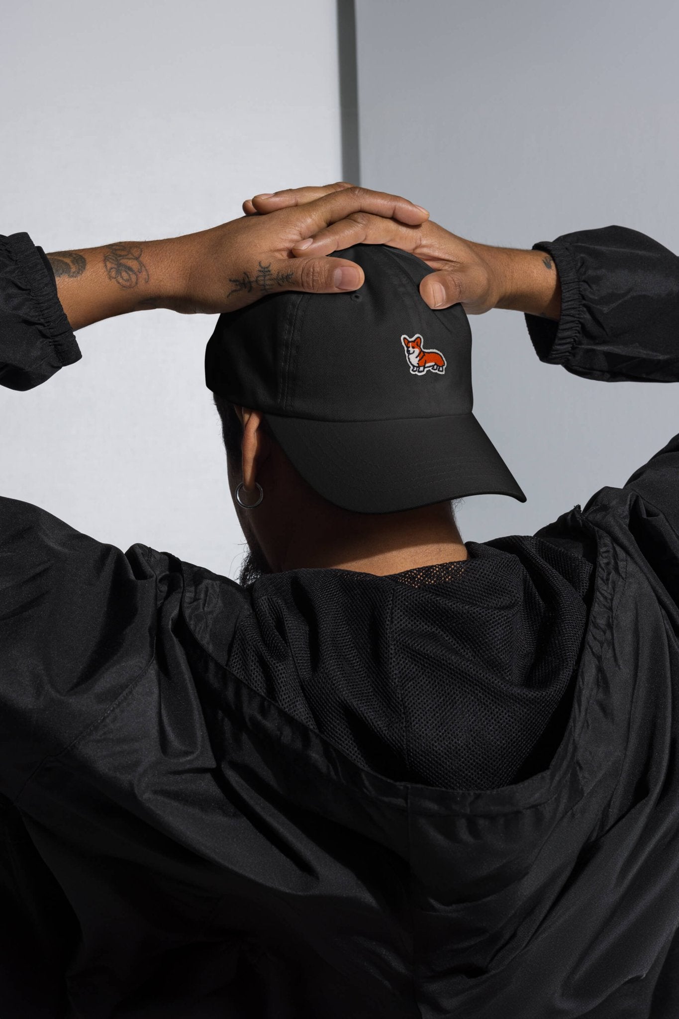 Wearing the Iconic Corgi Cap from Dogloverclothing.com, a person stands hands behind head, facing away. A small fox logo accents the black cap, while forearm tattoos add a unique edge to their Corgi-inspired style.