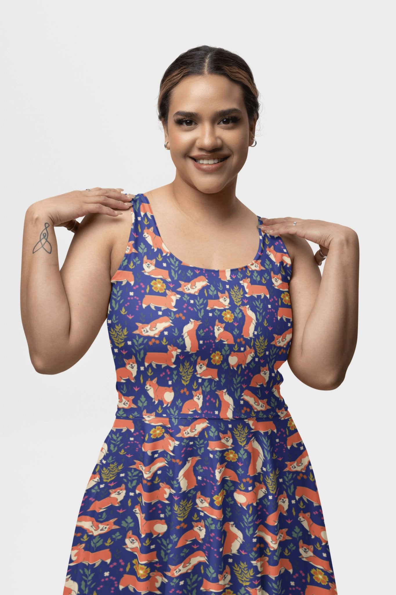 A woman joyfully poses with her hands near her shoulders, wearing a sleeveless dress adorned with colorful foxes and leaves on white. Her forearm reveals a small tattoo, capturing the vibrant whimsy of Dogloverclothing.coms Cute Corgi Garden Party Skater Dress.