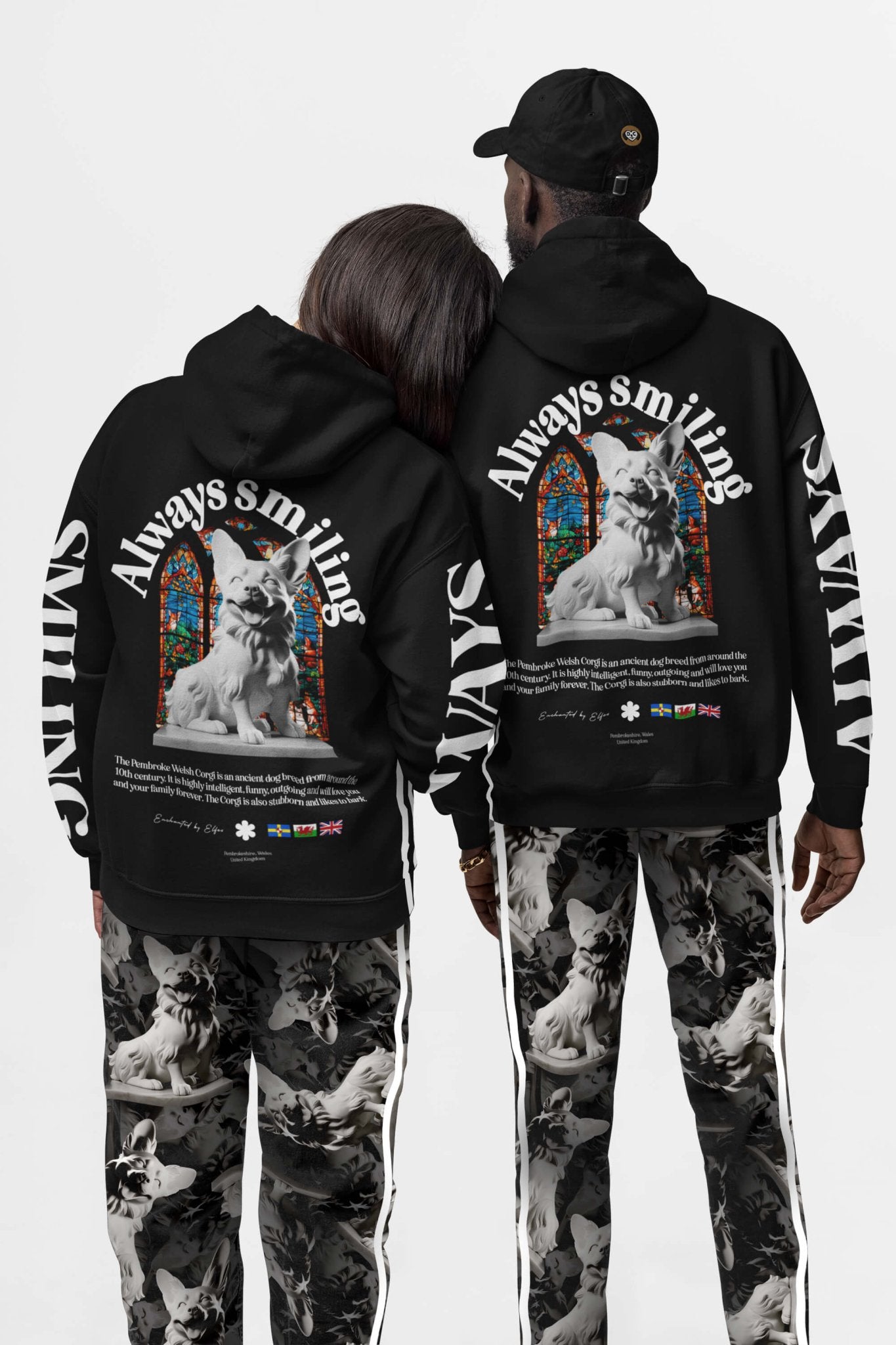 Two people are wearing Corgi Supreme Tribute Hoodies from Dogloverclothing.com, featuring a large statue graphic with Always Smiling text, stained glass images, and flags. Paired with patterned recycled material pants, they face away to showcase the eco-friendly design.