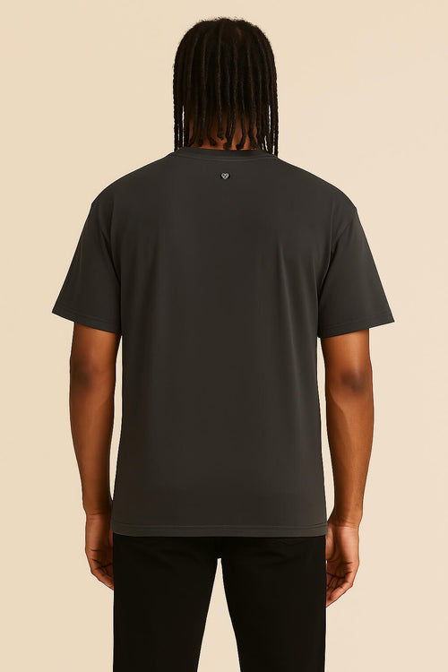 A person with medium-length dreadlocks is seen from the back, wearing the Oversized Dog Dad T-Shirt by Dogloverclothing.com in dark grey and black pants, standing against a beige background.