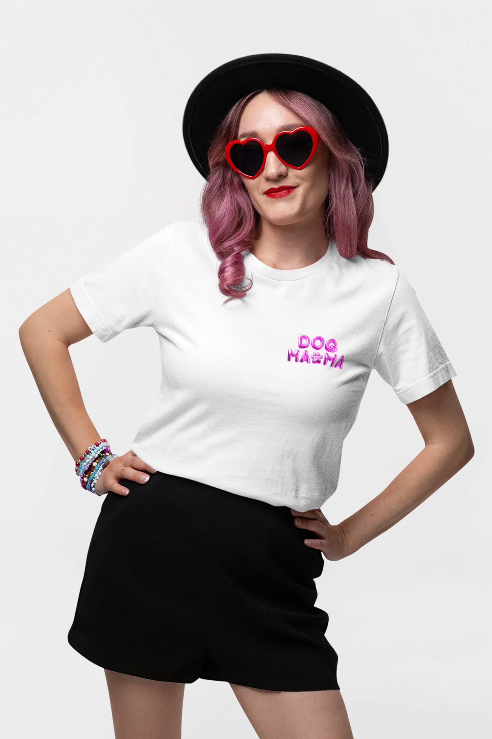 A person with pink hair, wearing a black hat and heart-shaped red sunglasses, confidently rocks a Dogloverclothing.com Ultra Soft Dog Mama Balloon T-Shirt. Black shorts complement the look as they pose with one hand on their hip, colorful bracelets adding flair to one wrist.