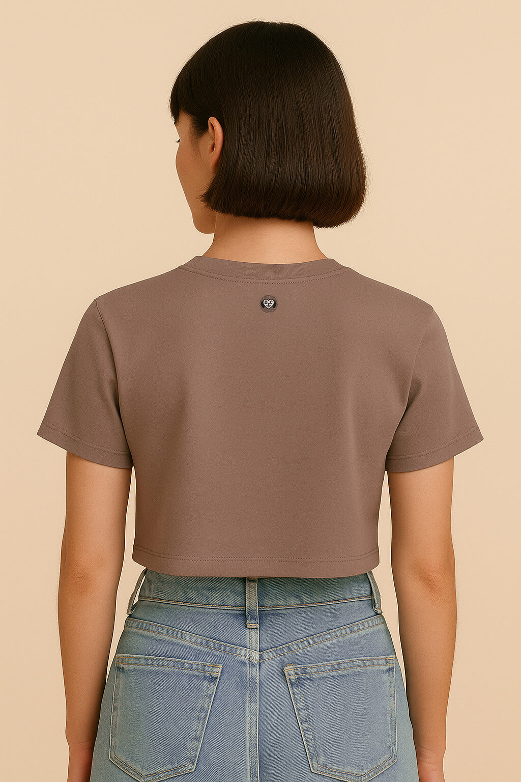 A person with short brown hair is shown from the back wearing the Oversized Dog Mom Crop Top by Dogloverclothing.com and high-waisted light blue jeans against a beige background, capturing an effortless Dog Mom Lifestyle vibe.