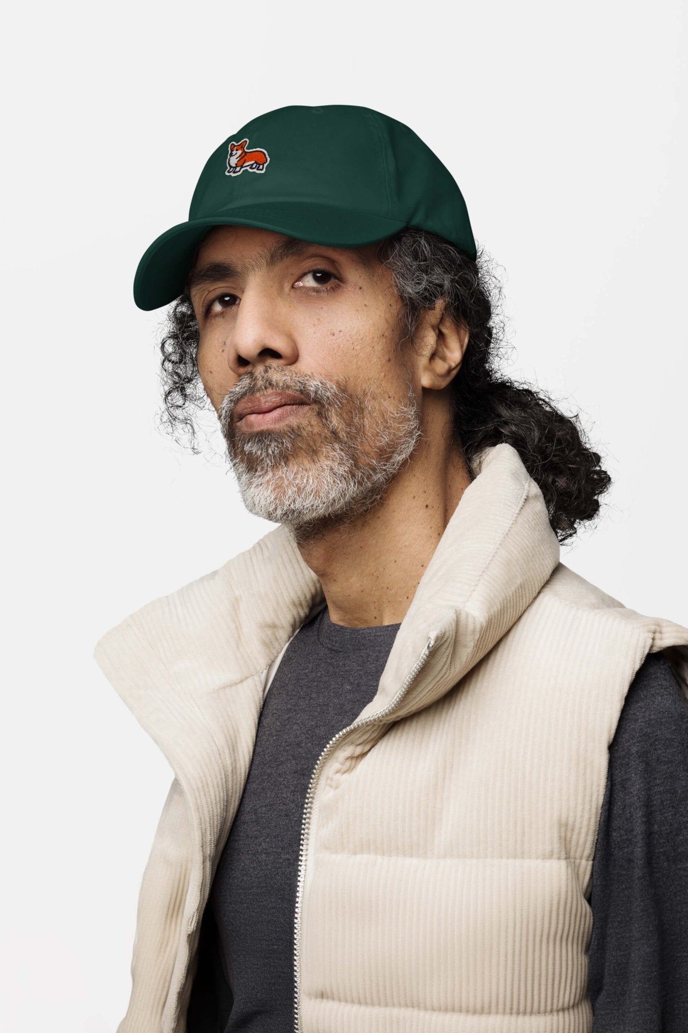 A man with curly gray hair and a beard wears the Iconic Corgi Cap by Dogloverclothing.com, featuring a playful dog logo, along with a beige vest and dark shirt, calmly looking at the camera against a plain light background.