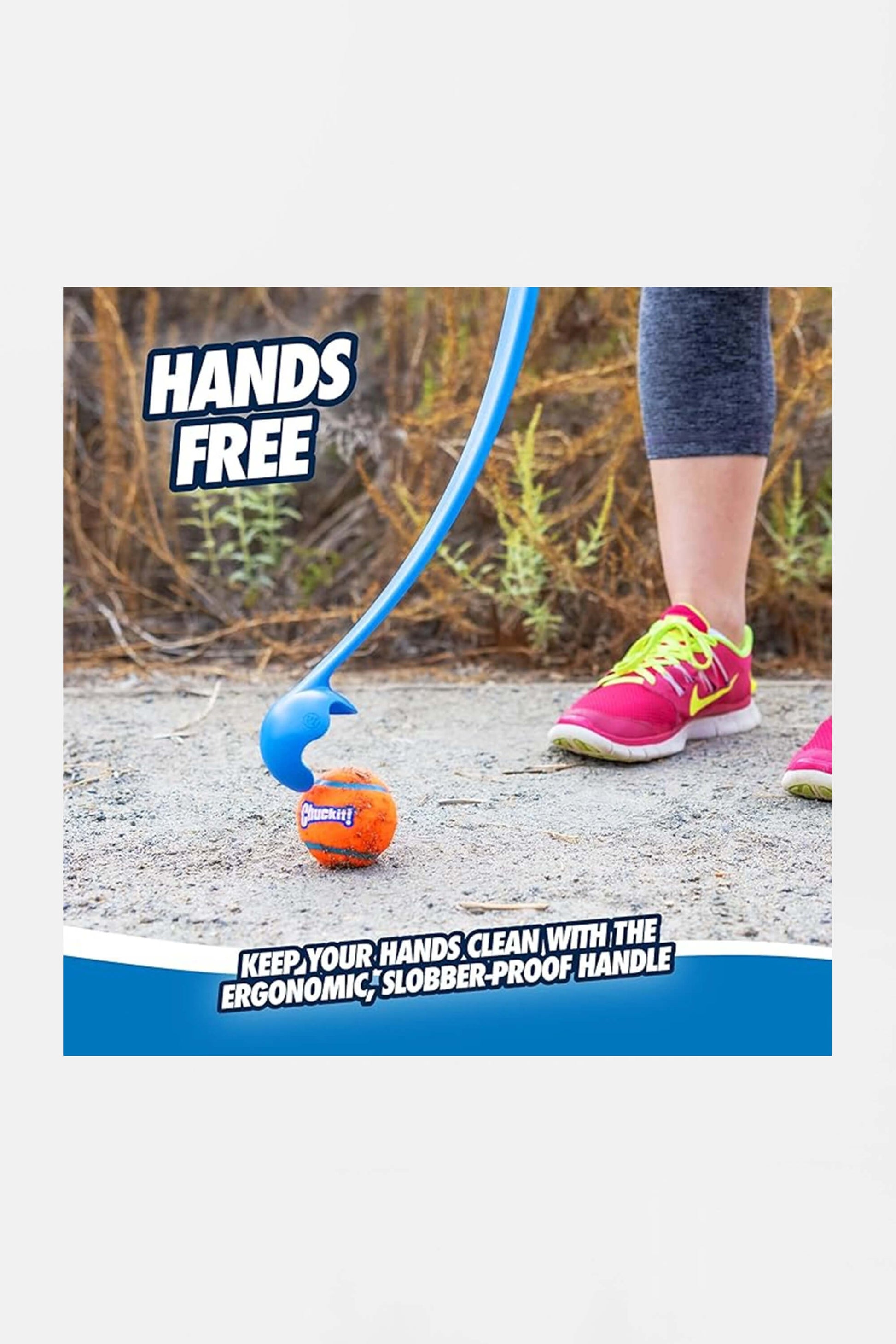 A person uses an Amazon Chuckit! 12 Dog Ball Launcher, picking up an orange and blue dog ball hands-free while wearing pink/yellow sneakers and gray leggings. The launcher has an ergonomic, slobber-proof handle for effortless fetch.