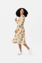A woman with curly hair smiles, standing on one leg, in a Cozy Corgi Long Sleeve Midi Dress from Dogloverclothing.com. The beige dress features playful orange cat patterns, blending comfort and style. She pairs it with white sneakers against a plain white backdrop.