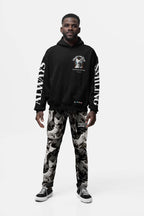 A man in a black Corgi Supreme Tribute Hoodie by Dogloverclothing.com with recycled material, black and gray patterned pants, and black sneakers stands against a white background, looking forward with his hands at his sides.