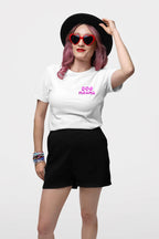 A person with pink hair poses confidently against a plain white background, wearing a black hat, heart-shaped red sunglasses, and an Ultra Soft Dog Mama Balloon T-Shirt by Dogloverclothing.com. Black shorts and colorful bracelets complete the look as they adjust their hat.