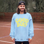A long-haired, bearded man on a sports court wears Dogloverclothing.coms blue Good Boy Sweater with GOOD BOY! in bold yellow letters. A backward black cap tops his head as lush greenery and wooden fences surround him, highlighting his love for dog-themed items.