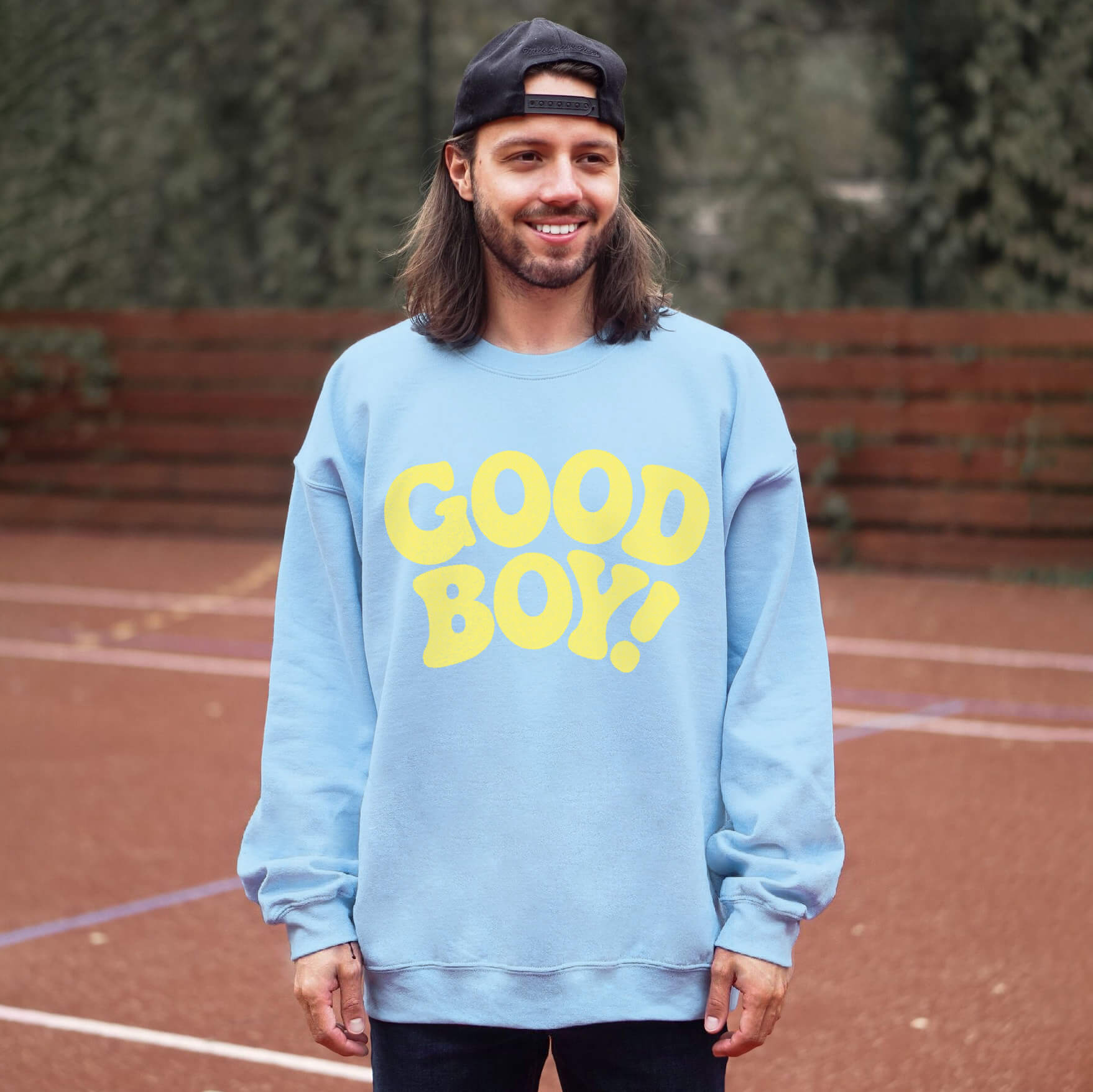 A long-haired, bearded man on a sports court wears Dogloverclothing.coms blue Good Boy Sweater with GOOD BOY! in bold yellow letters. A backward black cap tops his head as lush greenery and wooden fences surround him, highlighting his love for dog-themed items.