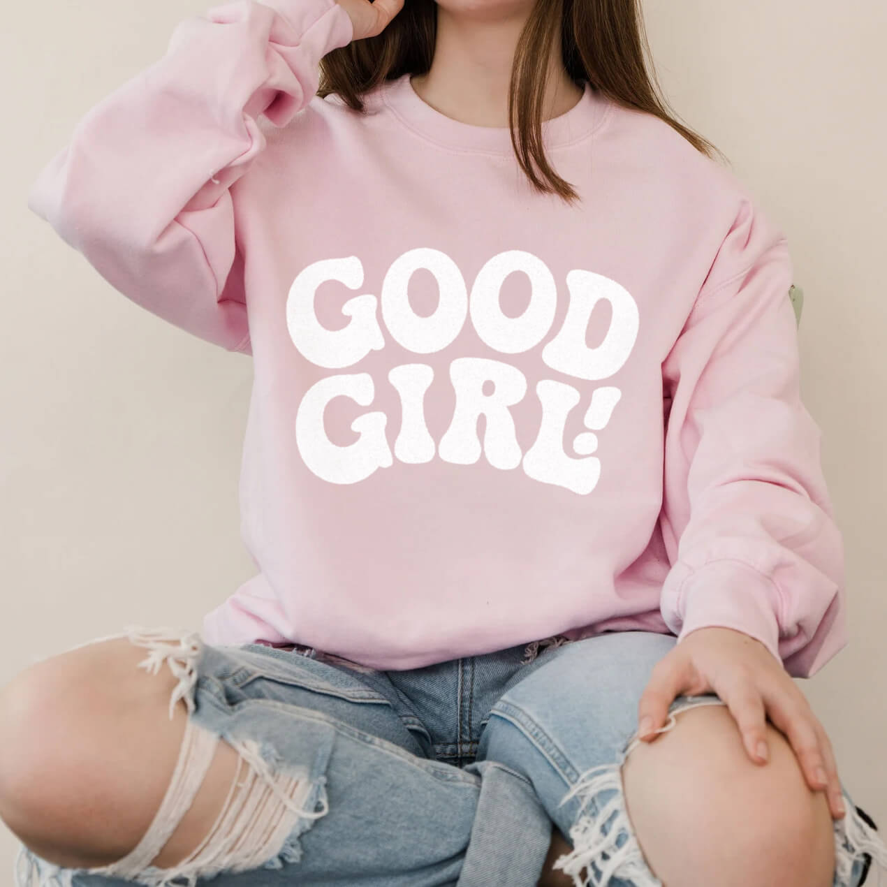 A person sits cross-legged, proudly wearing the Good Girl Sweatshirt from Dogloverclothing.com, showcasing their love for dog-themed items. Their ripped light blue jeans complete the look against a plain, neutral background.