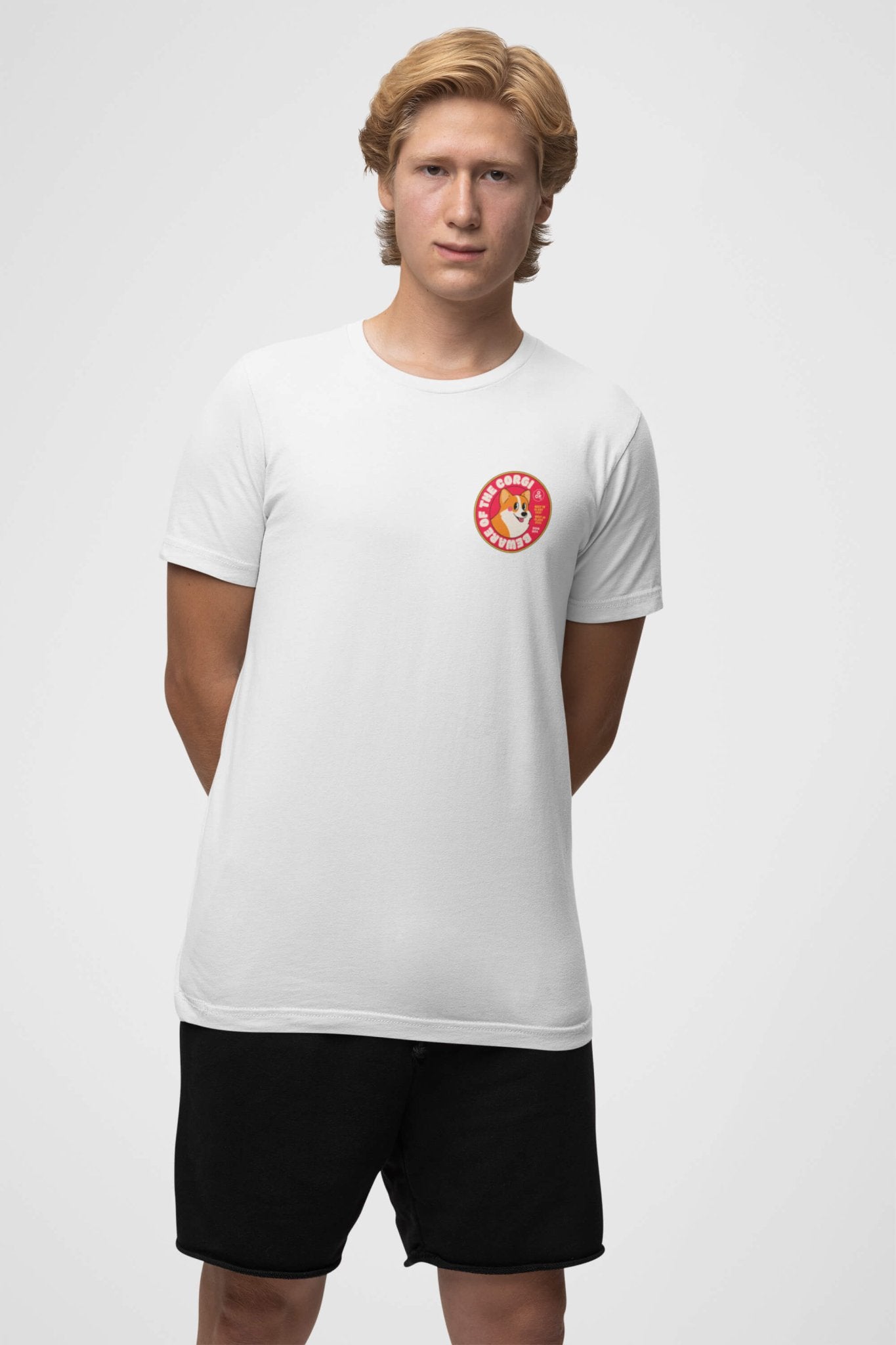 A young person stands against a plain backdrop in a Dogloverclothing.com Beware of the Corgi Supreme Unisex T-Shirt made of super comfy material, featuring a vibrant round Corgi life logo on the chest, paired with black shorts. Their arms are behind their back, exuding a casual and joyful vibe perfect for Corgi lovers.